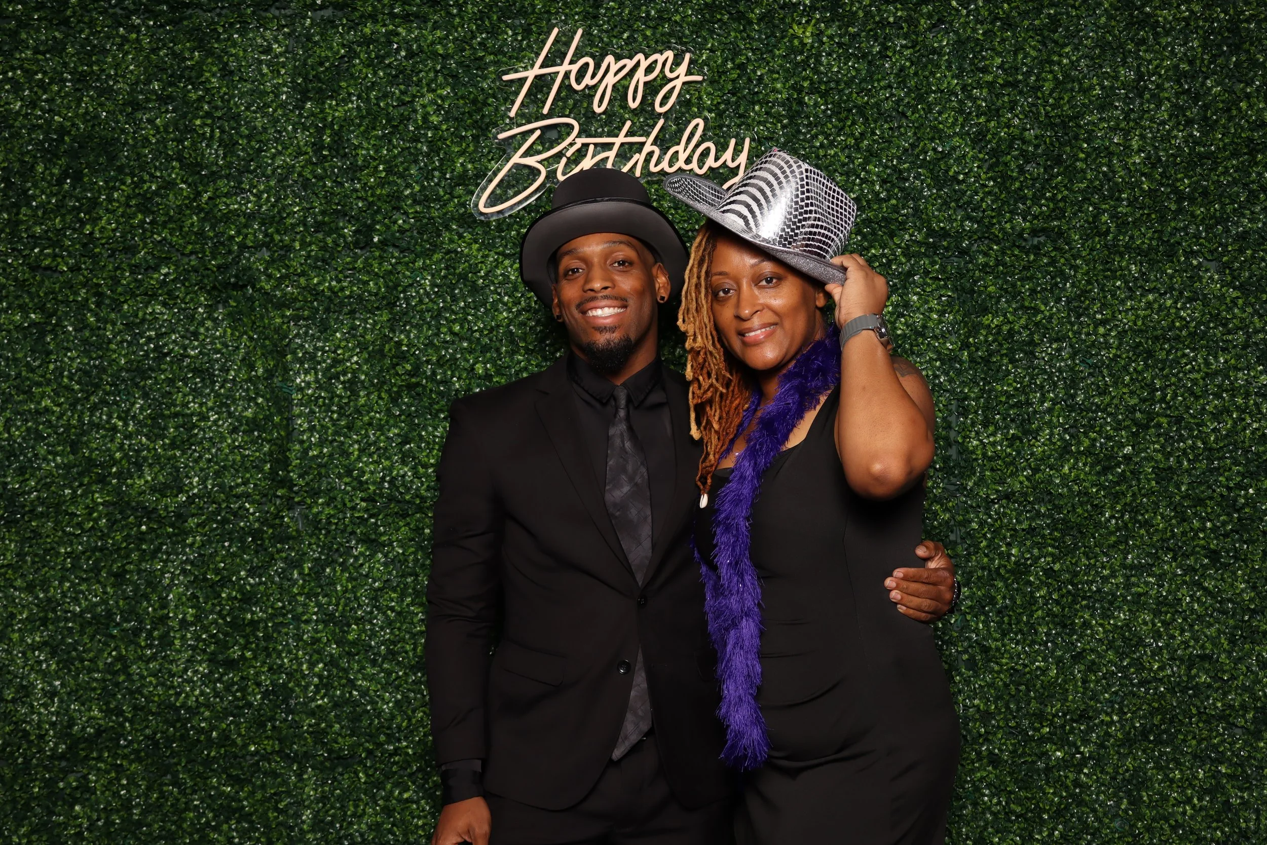 A man and woman dressed up with hats standing in front of a green hedge backdrop with illuminated "Happy Birthday" sign, smiling at the camera.