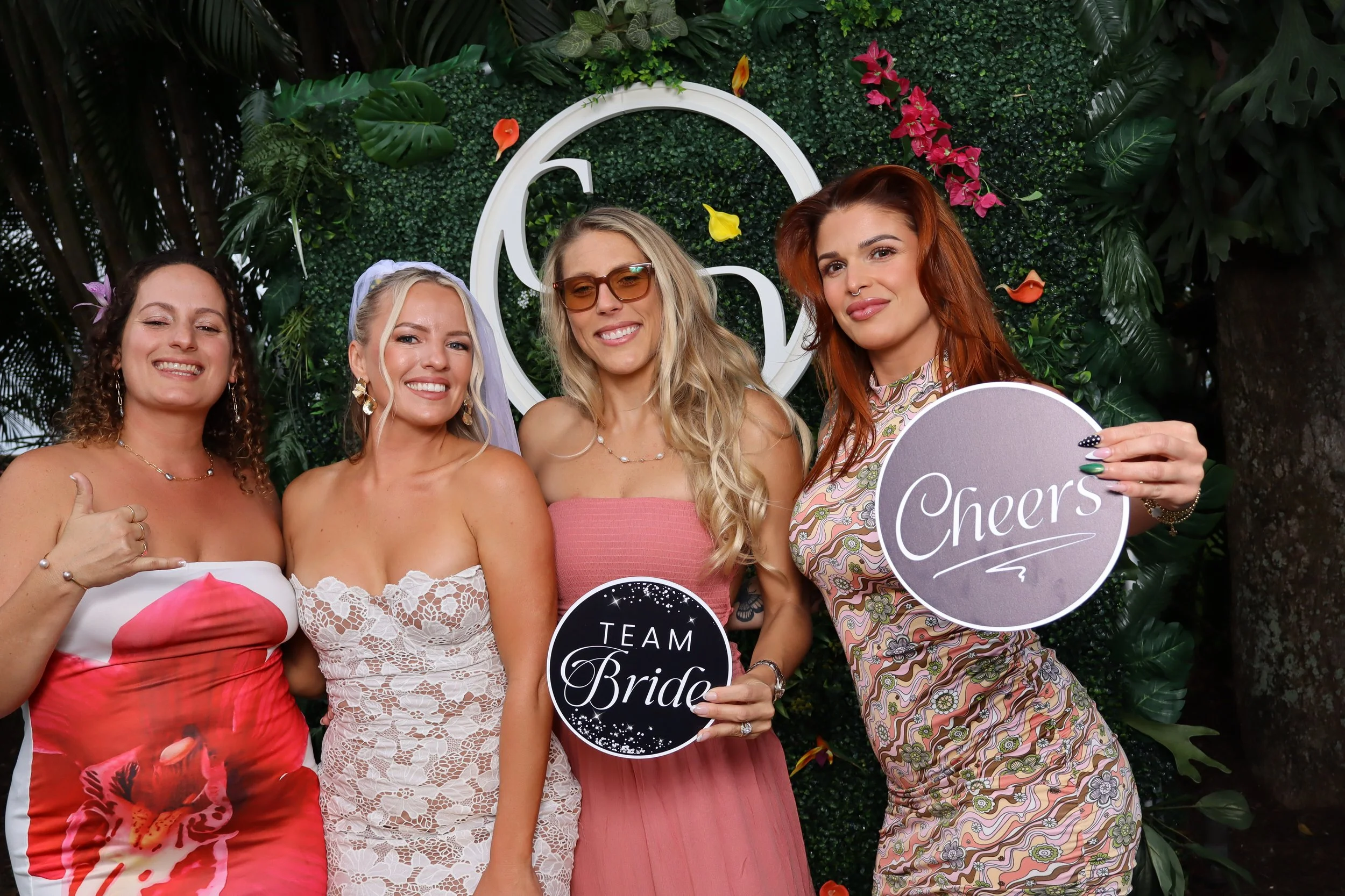Four women at a celebration party, smiling and holding signs that read "Team Bride" and "Cheers," standing in front of a lush, green decorative backdrop with flowers and a large circular design.