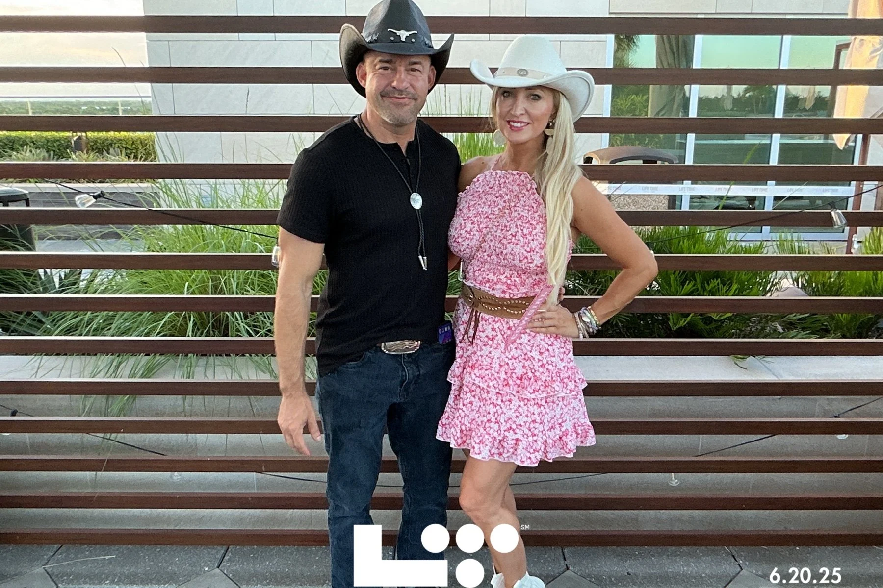 A man and woman wearing cowboy hats posing together outdoors in front of a wooden fence and green plants. The man is in a black shirt with a medallion necklace, and the woman is in a pink floral dress with a brown belt and white cowboy hat. The date 