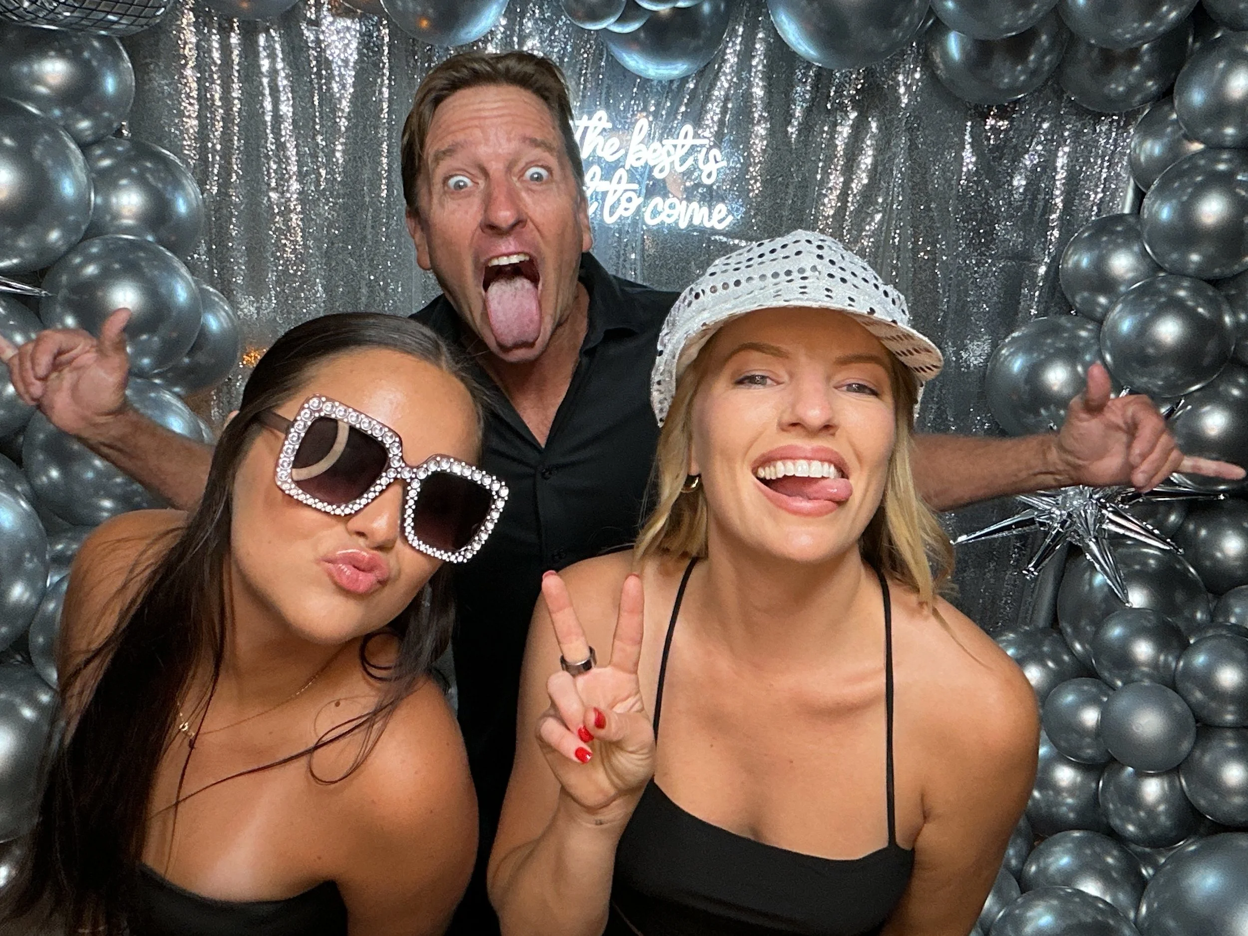 Three people posing at a celebratory event with metallic balloons and a sparkling backdrop. The woman on the left wears large, rhinestone sunglasses and makes a duck face. The woman on the right wears a white perforated hat, shows a peace sign, and s