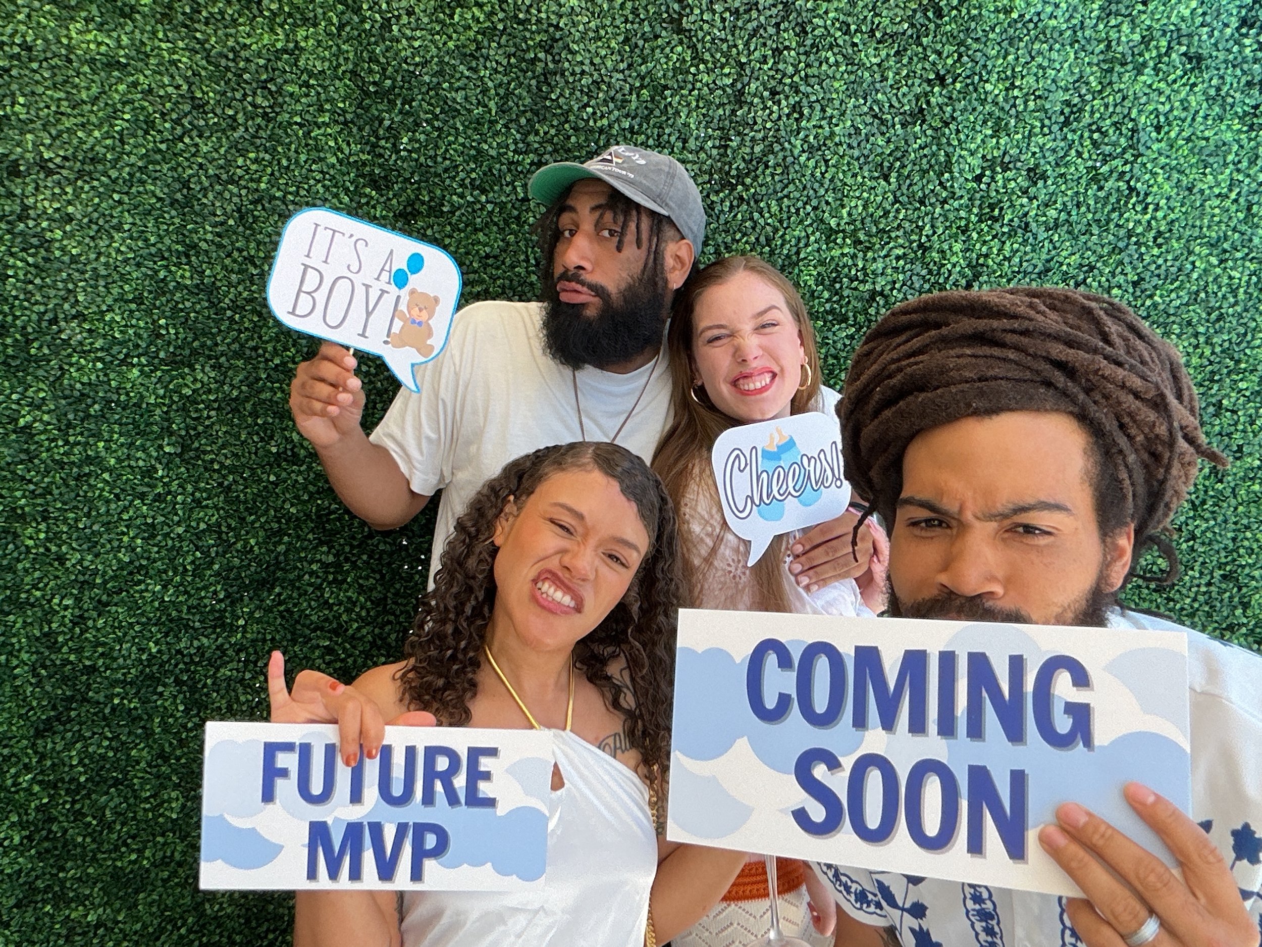 Four people standing in front of a green leafy background holding signs with celebratory messages. The person on the left is a woman holding a sign that says "FUTURE MVP". The second person is a man wearing a gray hat and holding a sign that says "IT