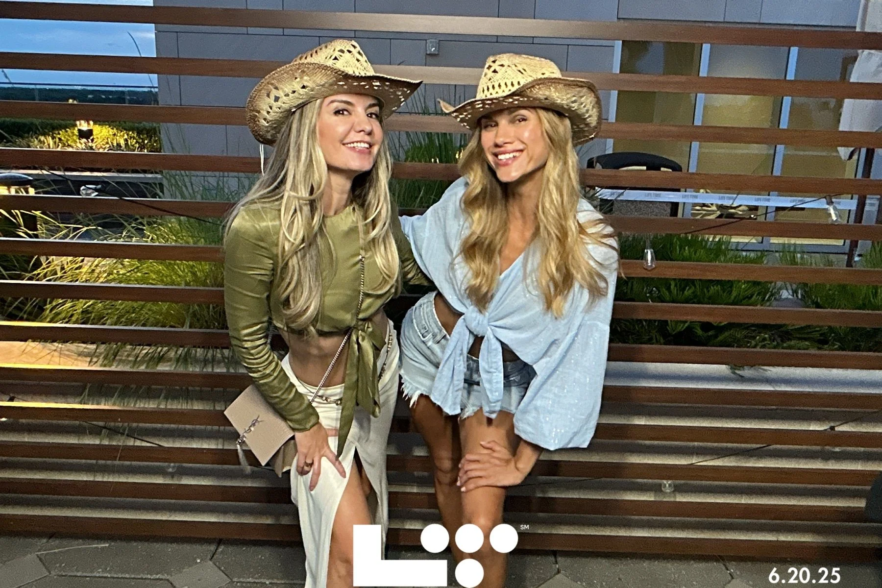 Two young women wearing cowboy hats smiling and posing together outdoors in front of a modern building and wooden fence, dressed in casual summer outfits.