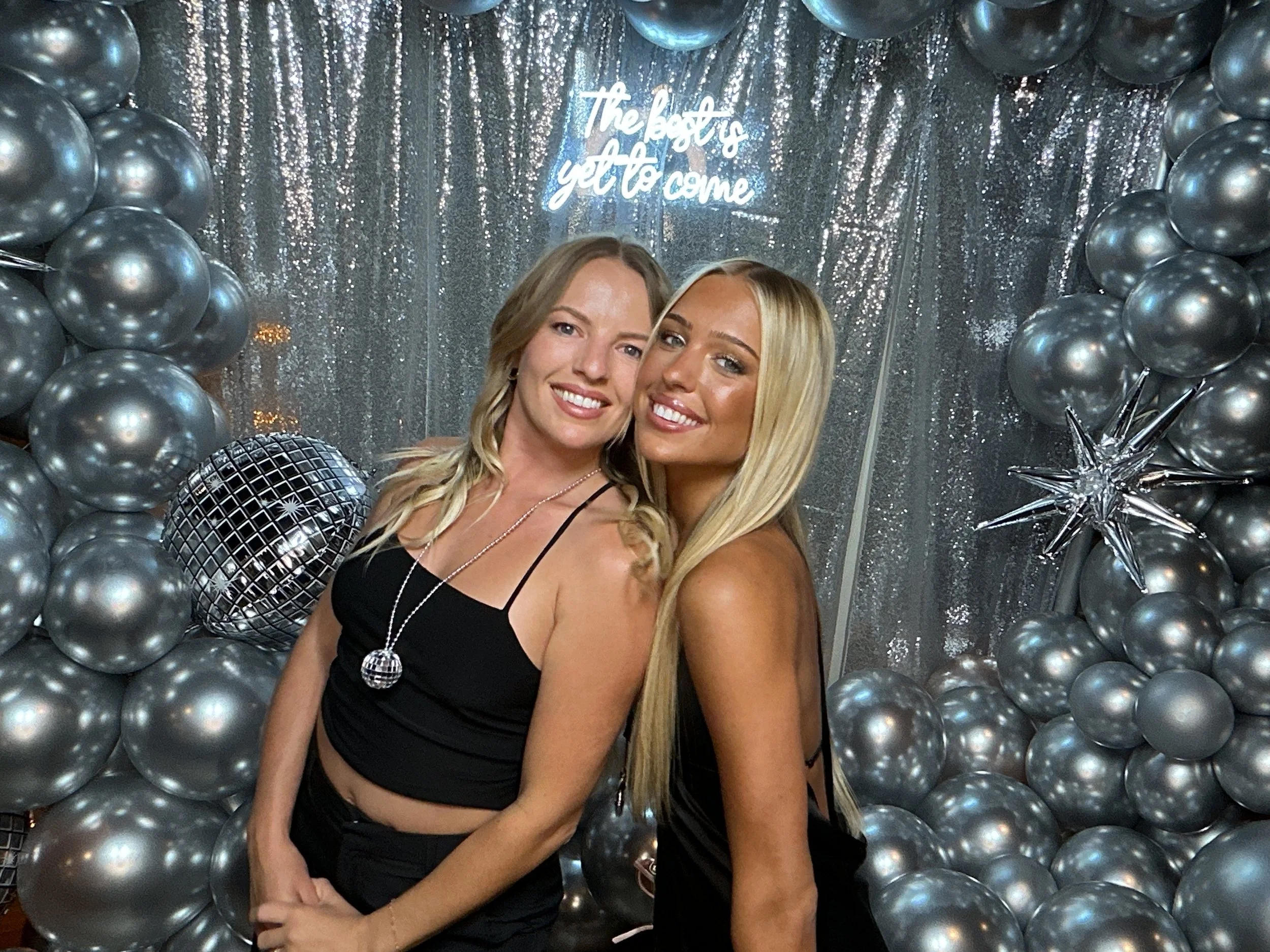 Two women smiling and posing at a party with a silver balloon backdrop, metallic star balloons, and a neon sign that says 'The best is yet to come' in the background.
