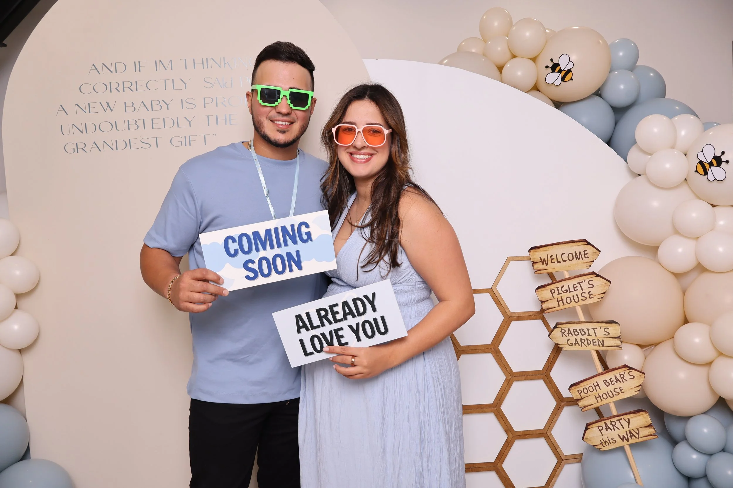 A man and woman smiling at a baby shower, holding signs that say "Coming Soon" and "Already Love You". The man wears green pixelated sunglasses, and the woman wears orange sunglasses. They are surrounded by cream and light blue balloons and decorativ