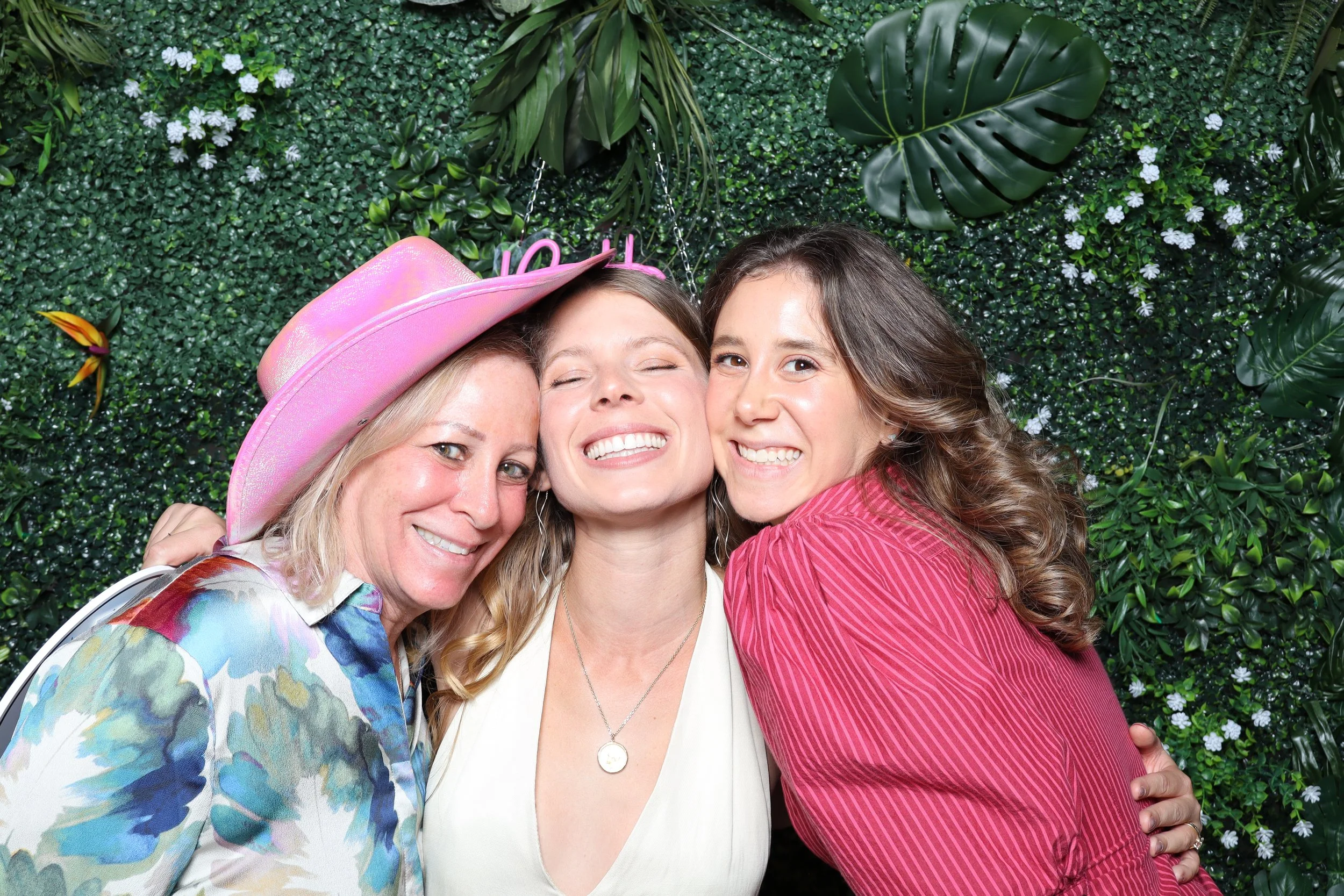 Three women smiling and hugging each other, standing in front of a green leafy wall with white flowers and large leaves, one wearing a pink hat.