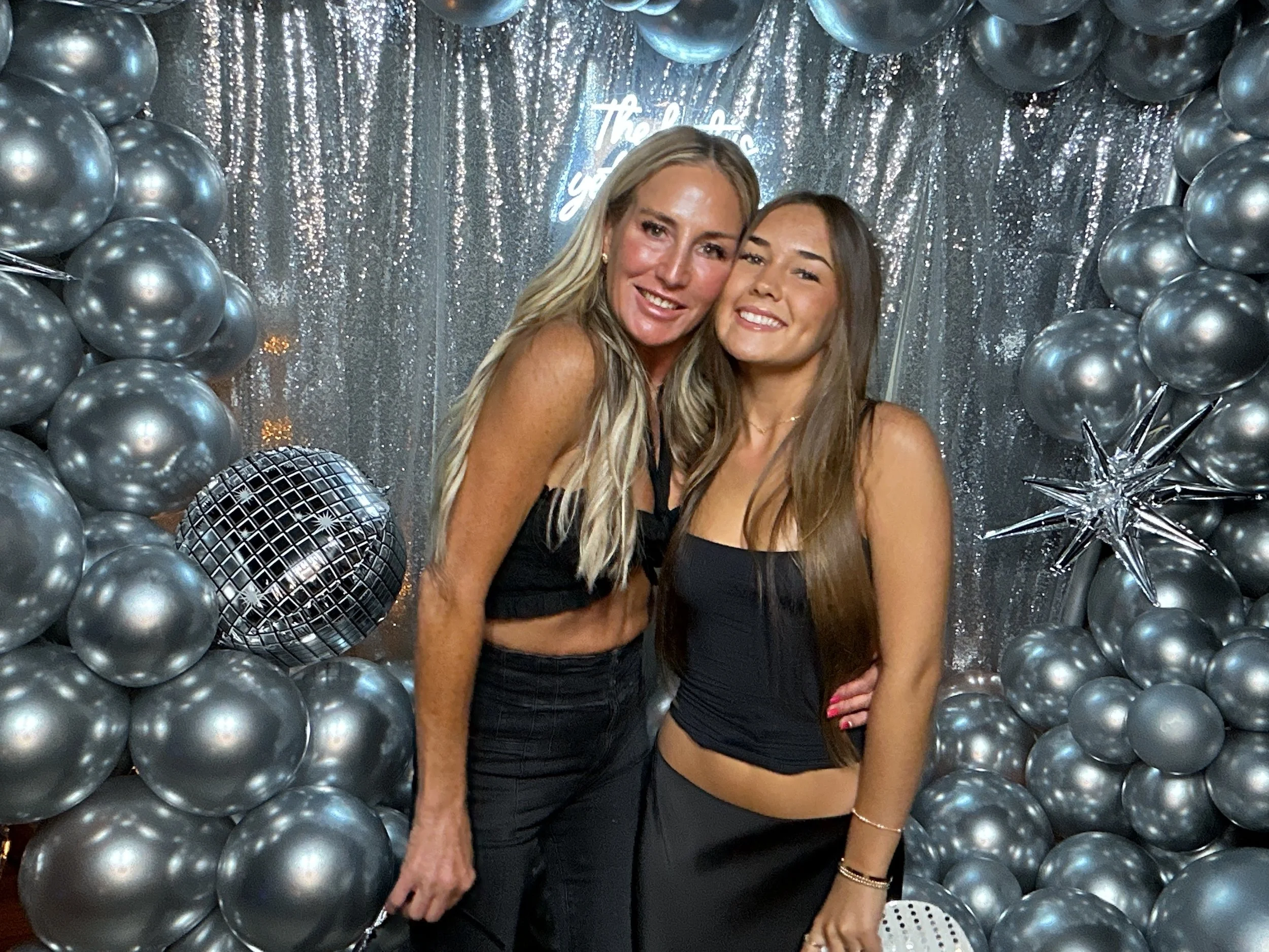 Two women posing together at a celebration decorated with silver balloons, a metallic disco ball, and a sparkly silver backdrop. They are smiling, dressed in black, and the scene suggests a party or special event.