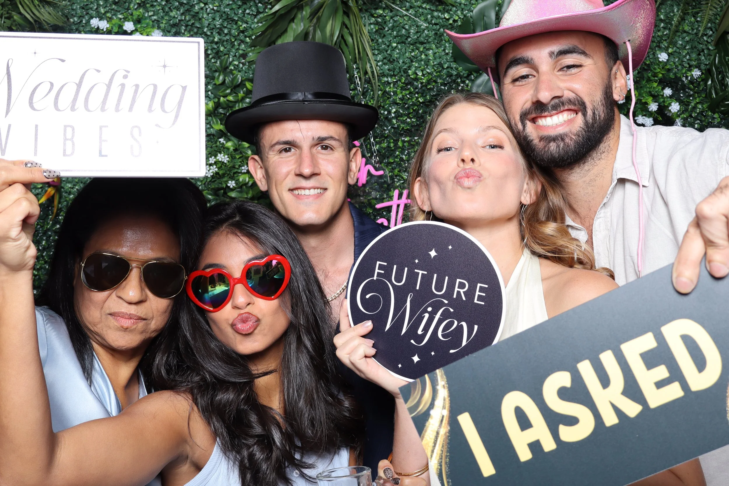 A group of six friends at a wedding celebration holding props and signs, including one that says 'Future Wife' and another that says 'I Asked.' They are dressed casually with some wearing hats and sunglasses, and they are posing against a green leafy