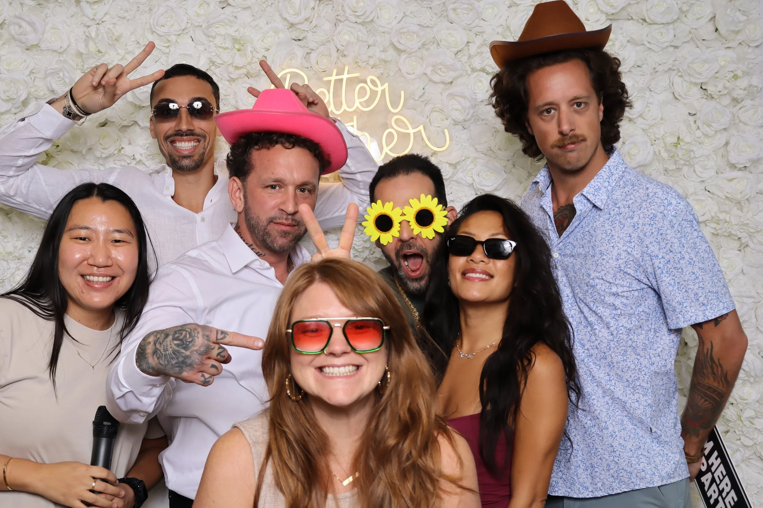 Group of people having fun at a photo booth with a white floral backdrop and a neon sign that says 'Better Together.' They are wearing playful accessories, including pink and brown hats, sunglasses, and sunflower and bunny ears, smiling and posing fo