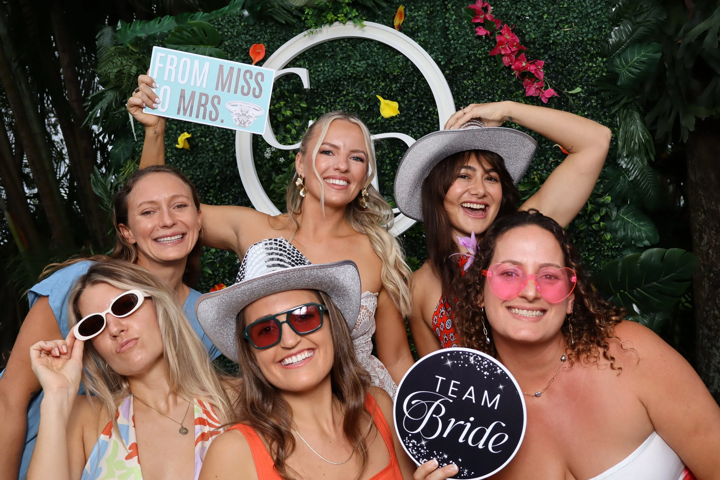 Group of women celebrating at a bridal shower with various playful accessories, holding signs that say 'From Miss to Mrs.' and 'Team Bride', in front of a floral and greenery backdrop.