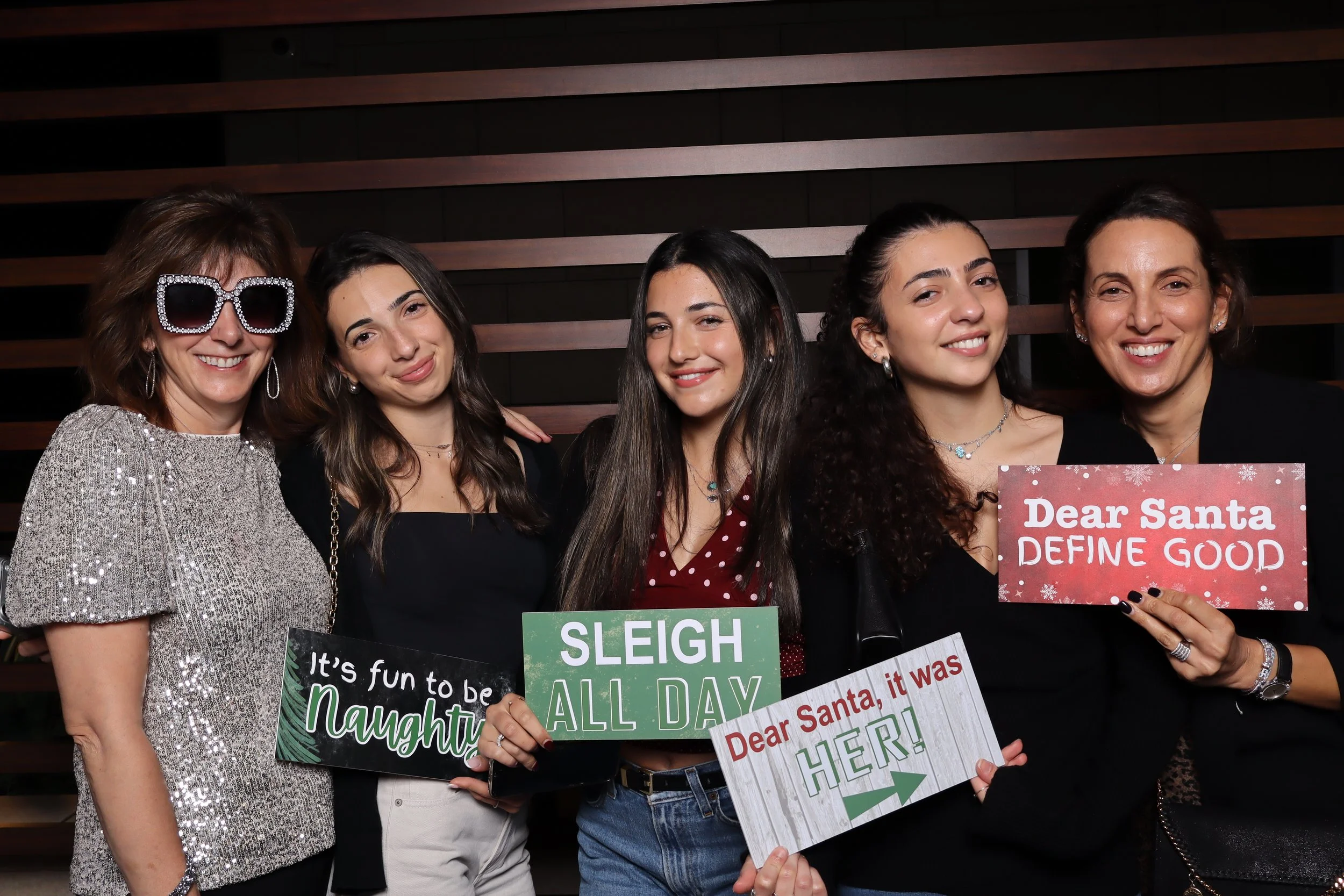 Five women smiling at a holiday party, holding signs that read 'It's fun to be Naughty,' 'Sleigh All Day,' 'Dear Santa, it was HER!,' and 'Dear Santa, DEFINE GOOD,' against a wooden wall background.