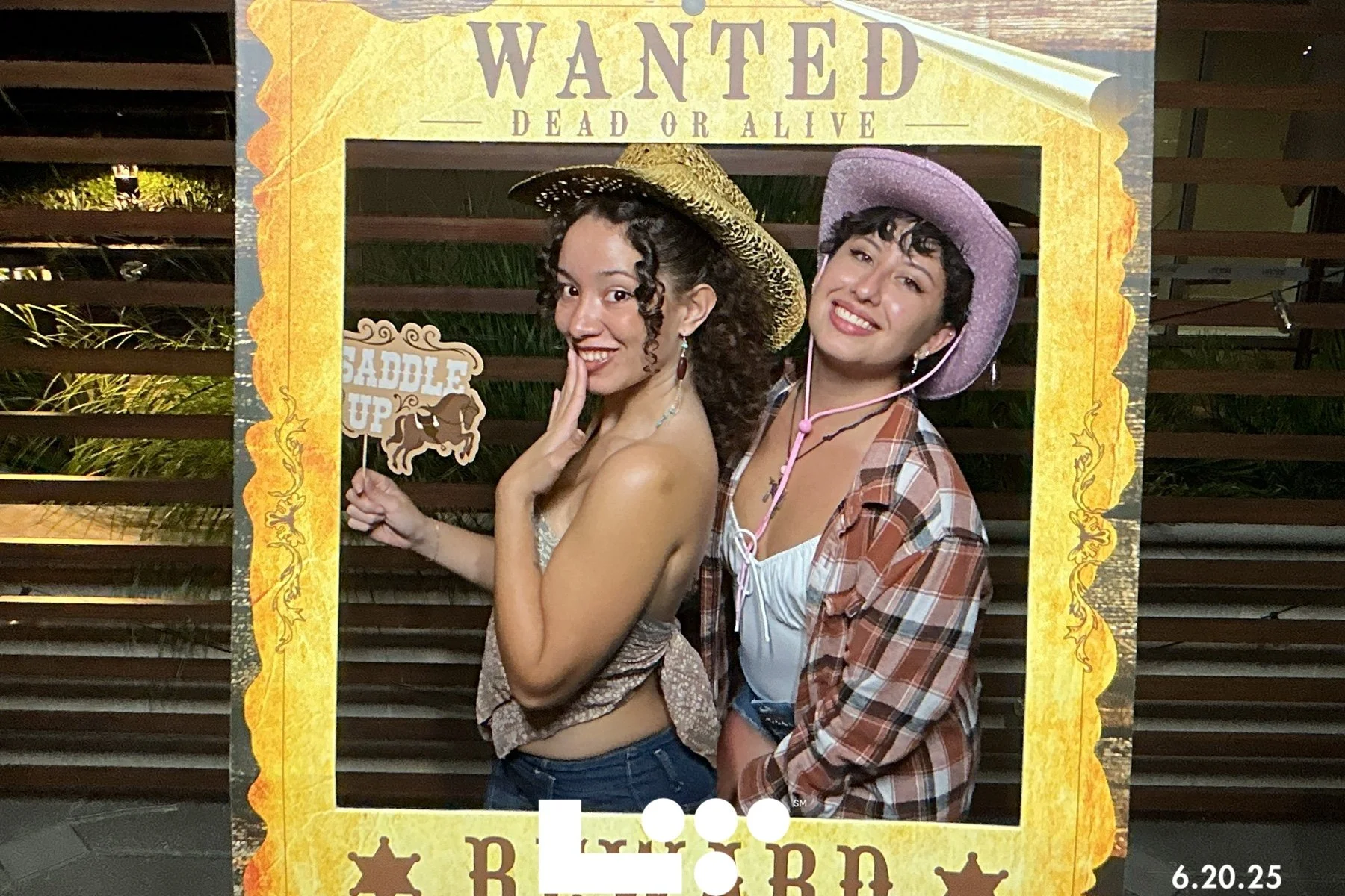 Two young women dressed in country or western costumes posing behind a decorative frame that says 'Wanted Dead or Alive' and 'Rodeo.' One woman is holding a sign that says 'Saddle Up,' and both are wearing cowboy hats.