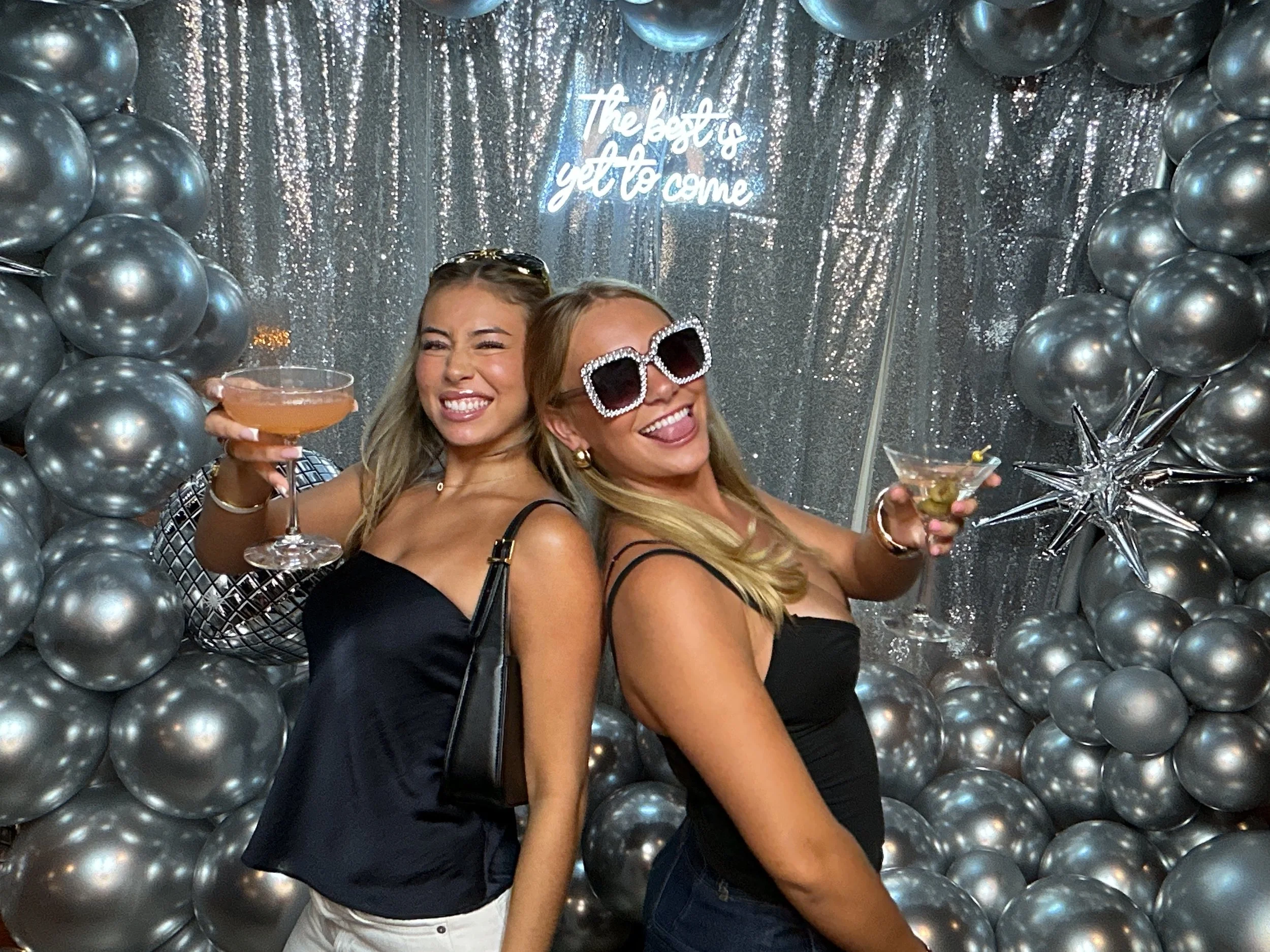 Two women at a celebration, posing with drinks in front of a backdrop with silver balloons and a shiny silver curtain. One woman wears large sunglasses and is making a playful face, while the other holds a cocktail and smiles. The backdrop has a sign
