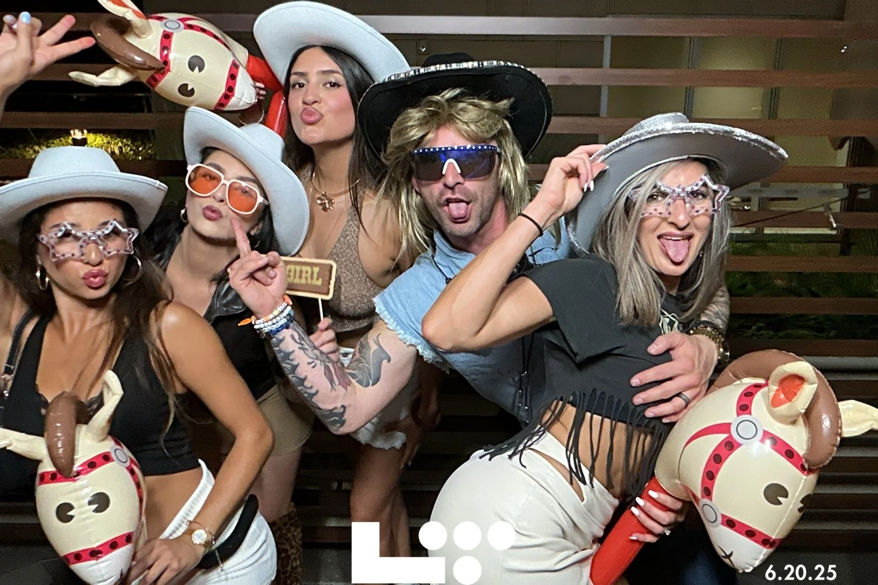 Group of five women and one man celebrating at a party, wearing large white cowboy hats, star-shaped sunglasses, and holding inflatable horses. The women are making playful faces, puckering their lips and sticking out tongues, while the man is sticki
