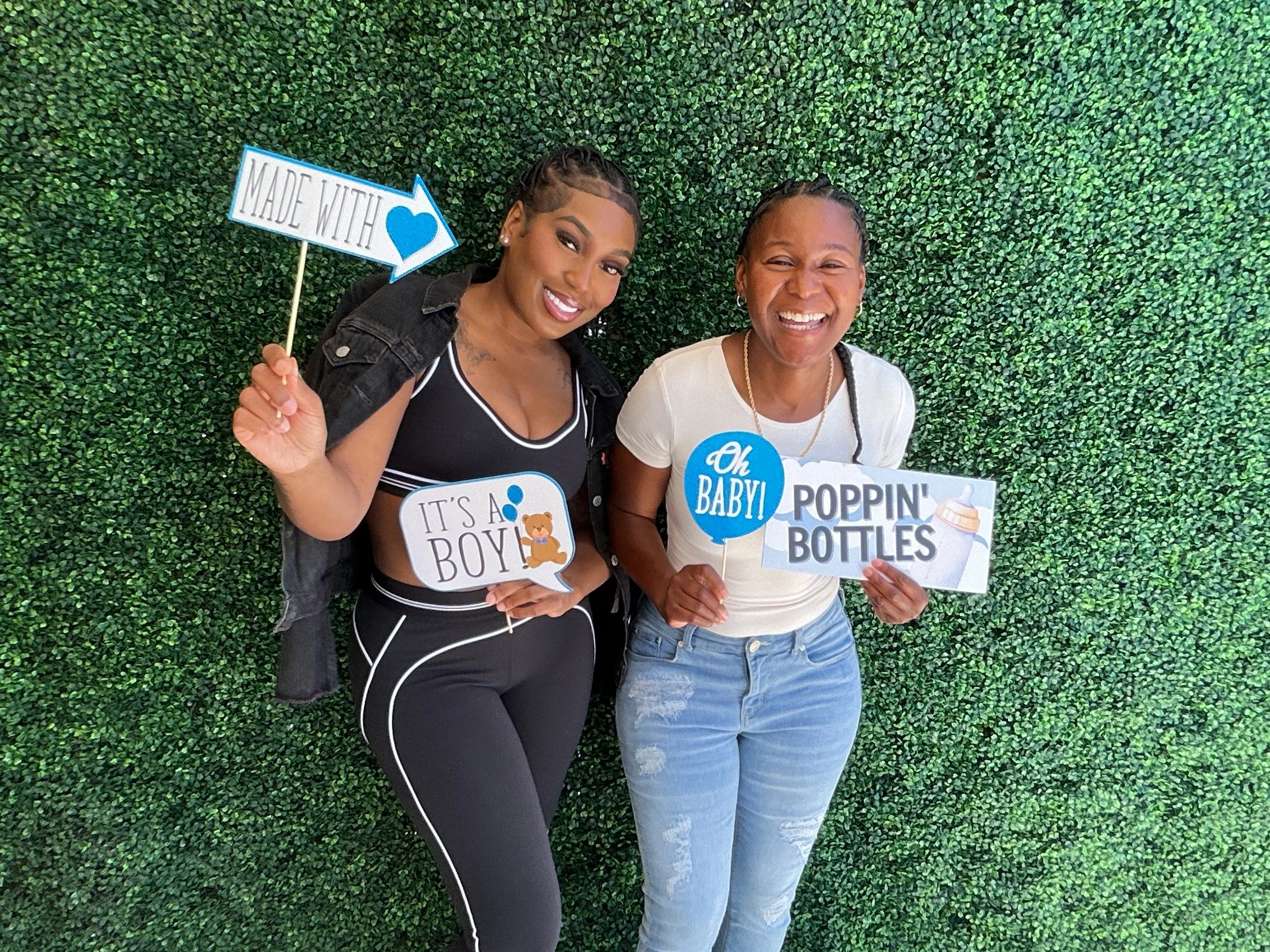 Two women standing in front of a green leafy wall, smiling and holding baby shower signs. One sign says 'Made with ♥', another says 'It's a Boy!', the other 'Oh Baby!', and 'Poppin Bottles' with an illustration of a baby bottle.