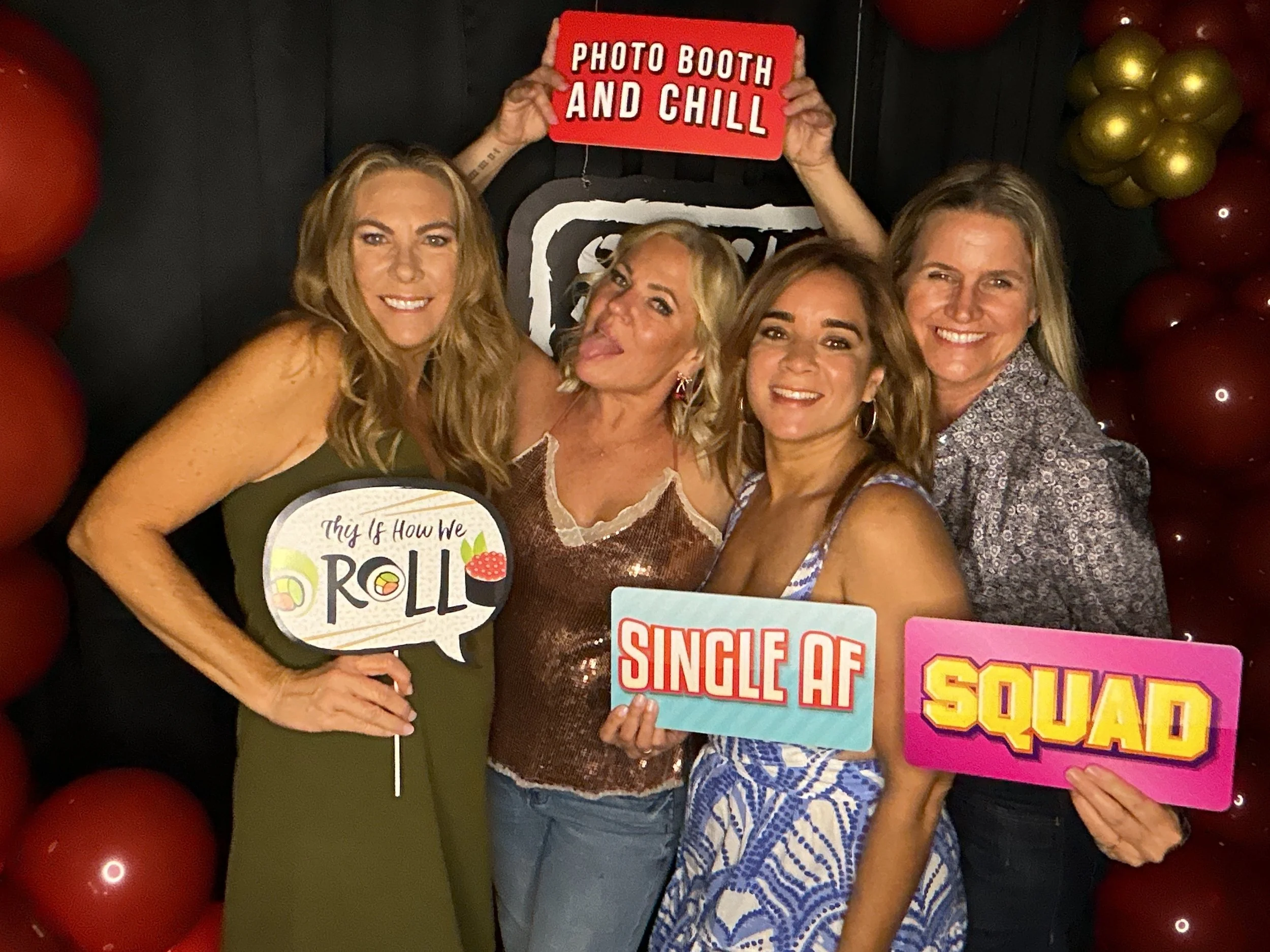 Four women posing together at a party, holding colorful signs with fun phrases. One sign reads 'This is how we roll,' another reads 'Single AF,' the third 'Squad,' and the last 'Photo booth and chill.' They are standing in front of a backdrop decorat