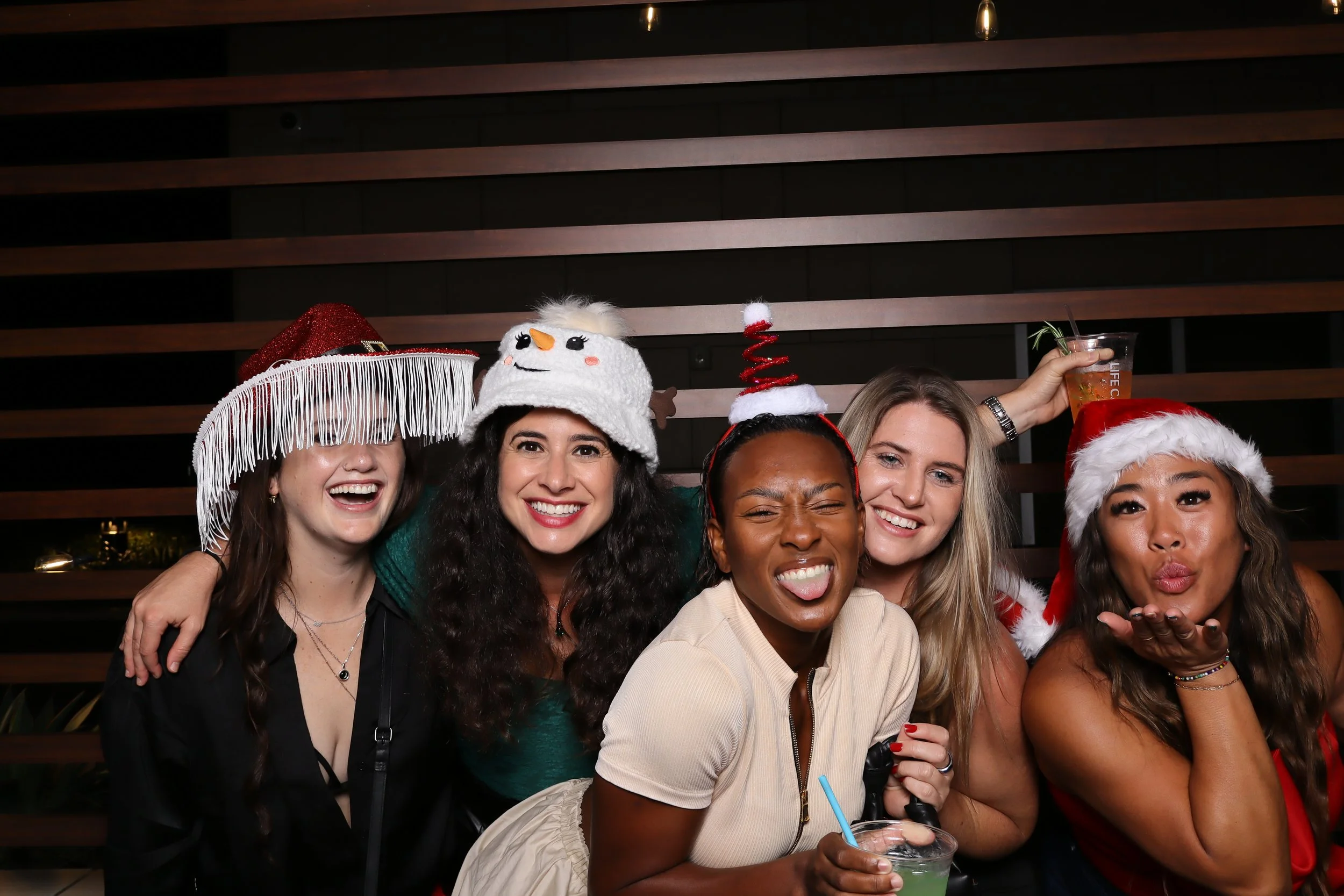 Group of five women celebrating at a holiday party, wearing festive hats including Santa hats and Christmas-themed accessories, holding drinks, and smiling for the camera.