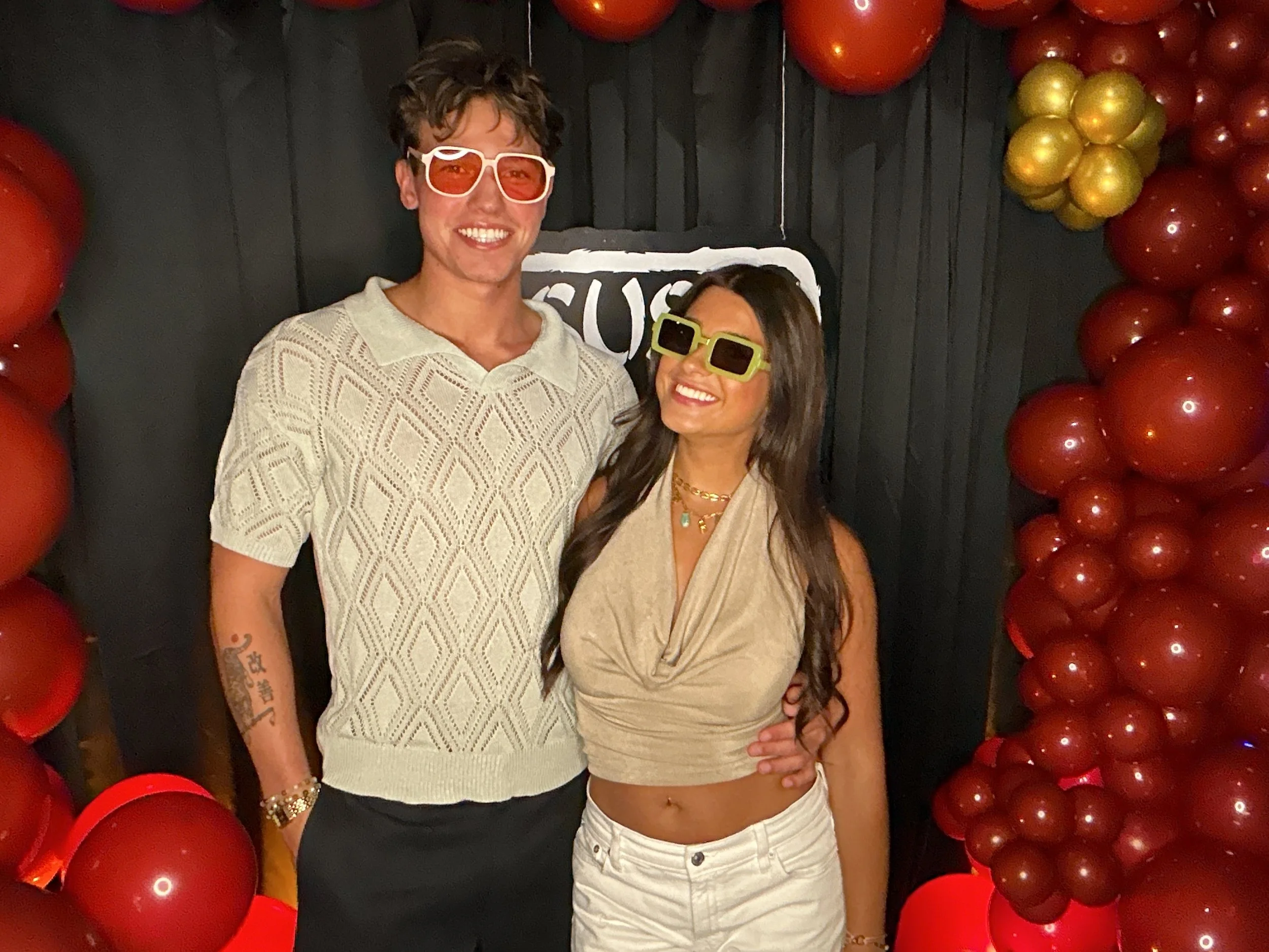 A man and woman wearing sunglasses smiling at a photo booth with red and gold balloons surrounding them.