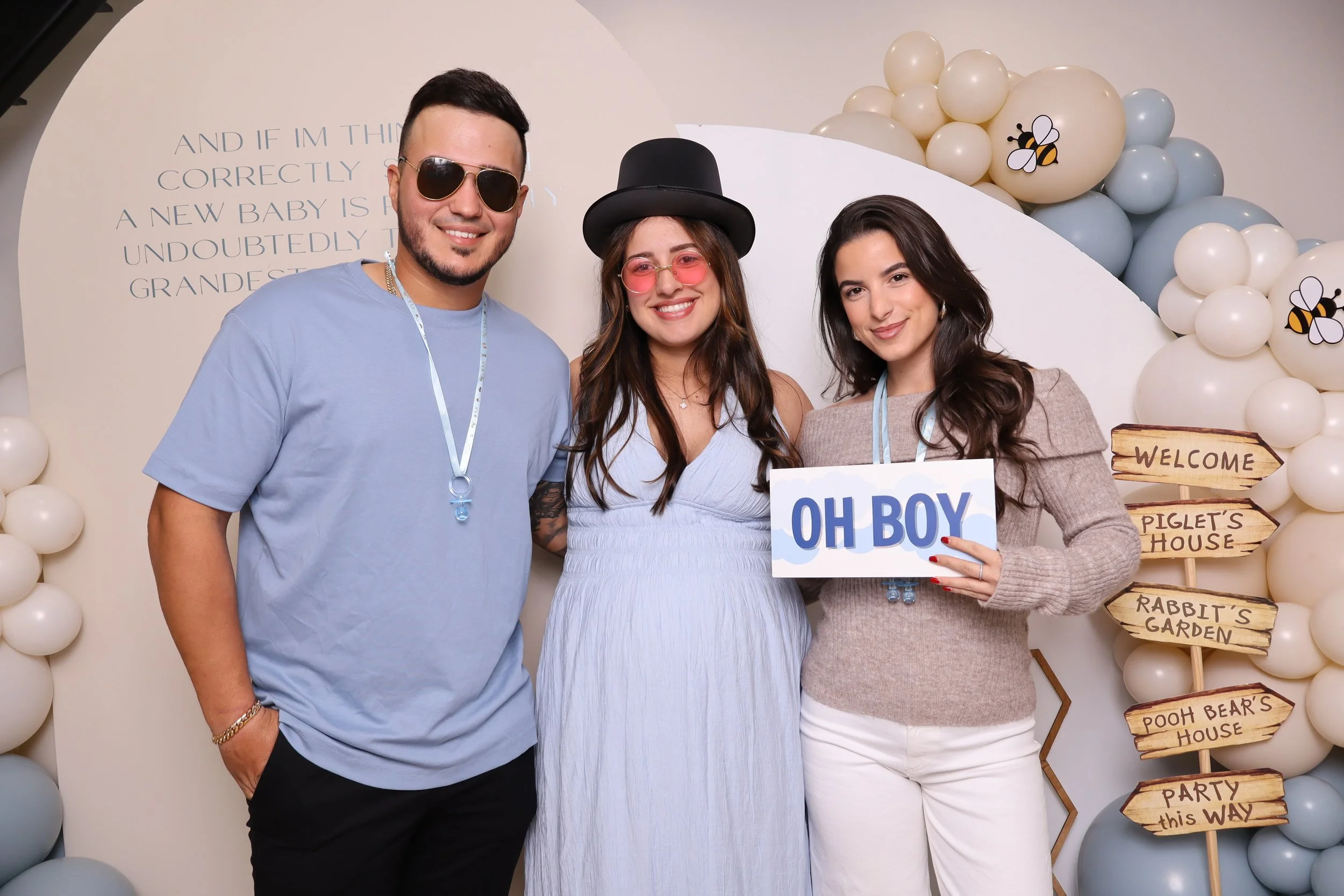 A group of three people celebrating a gender reveal party, with a backdrop of white and light blue balloons, some with bee illustrations, and a sign that reads 'OH BOY'. One woman wears a top hat and pink-tinted sunglasses, holding a sign that says '