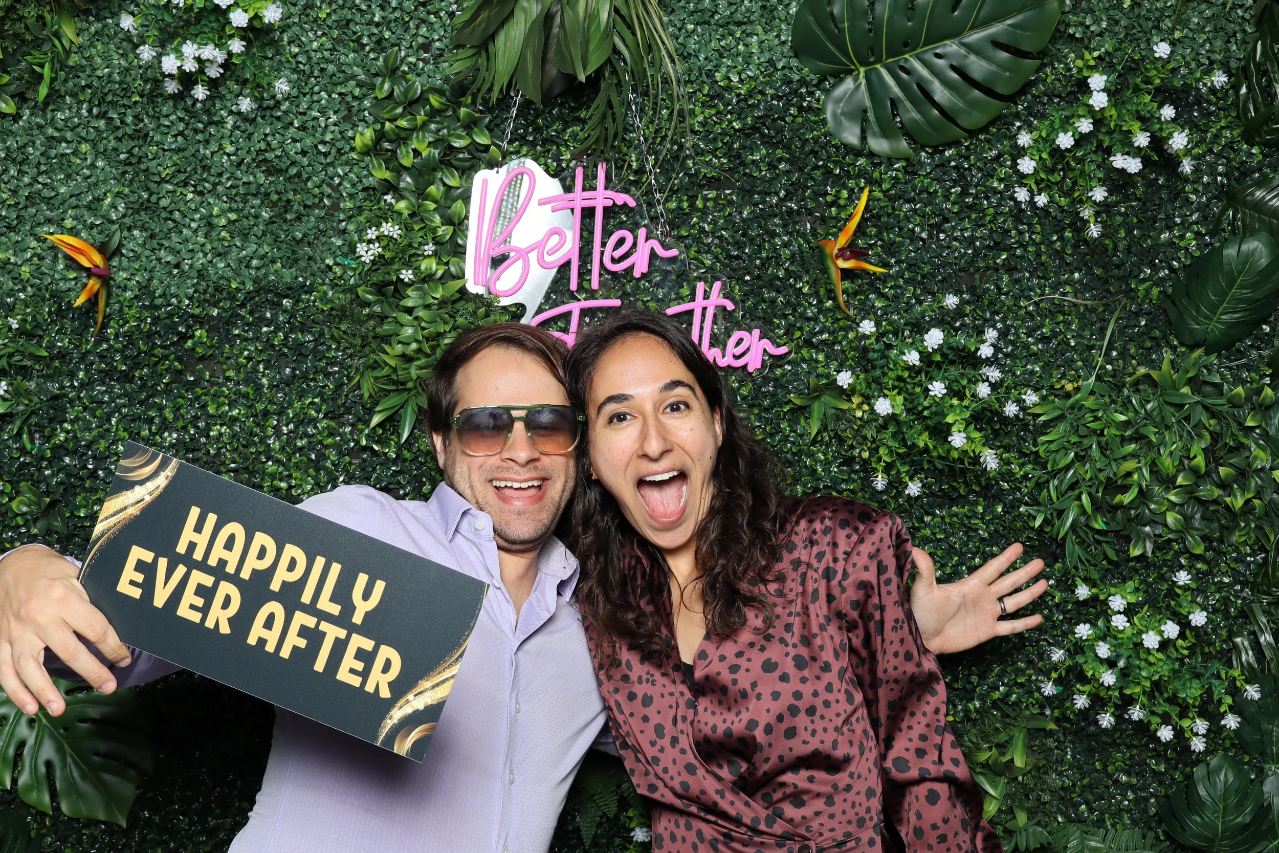 A man and woman celebrating in front of a green leafy wall with a pink neon sign reading 'Better Together'. The man is wearing sunglasses and a light purple shirt, holding a sign that says 'Happily Ever After'. They are smiling and appearing joyful.