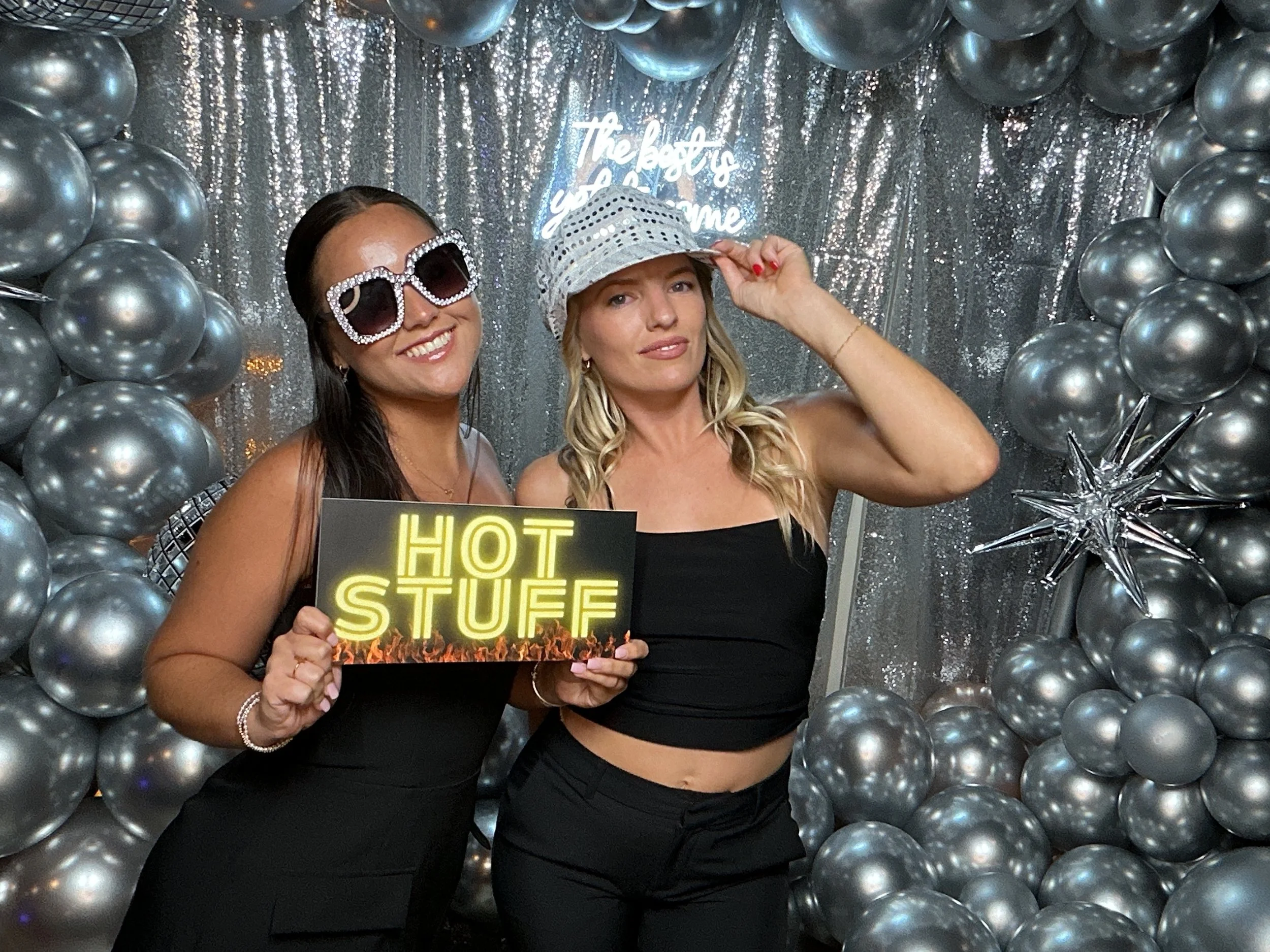 Two women at a party holding a sign that says 'HOT STUFF', standing in front of a silver balloon backdrop with a neon sign. The woman on the left is wearing large sunglasses and a black dress. The woman on the right is wearing a silver hat and a blac