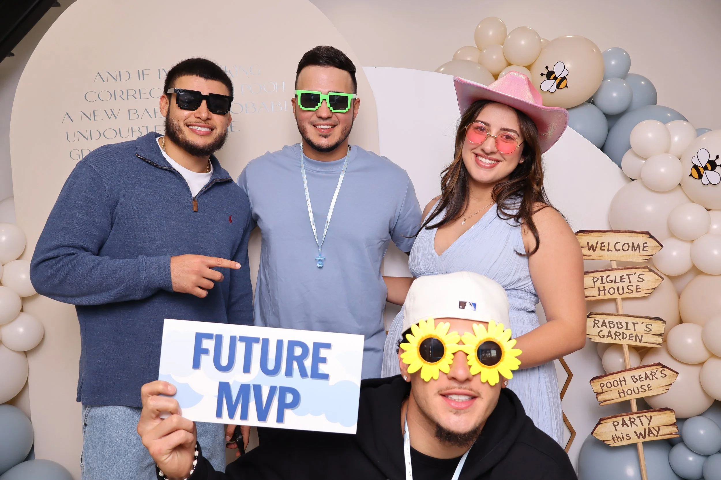 A gathering of people on a beach with a blue sky, some are seated on chairs, others are walking or standing near the shoreline, with umbrellas and boats in the distance.