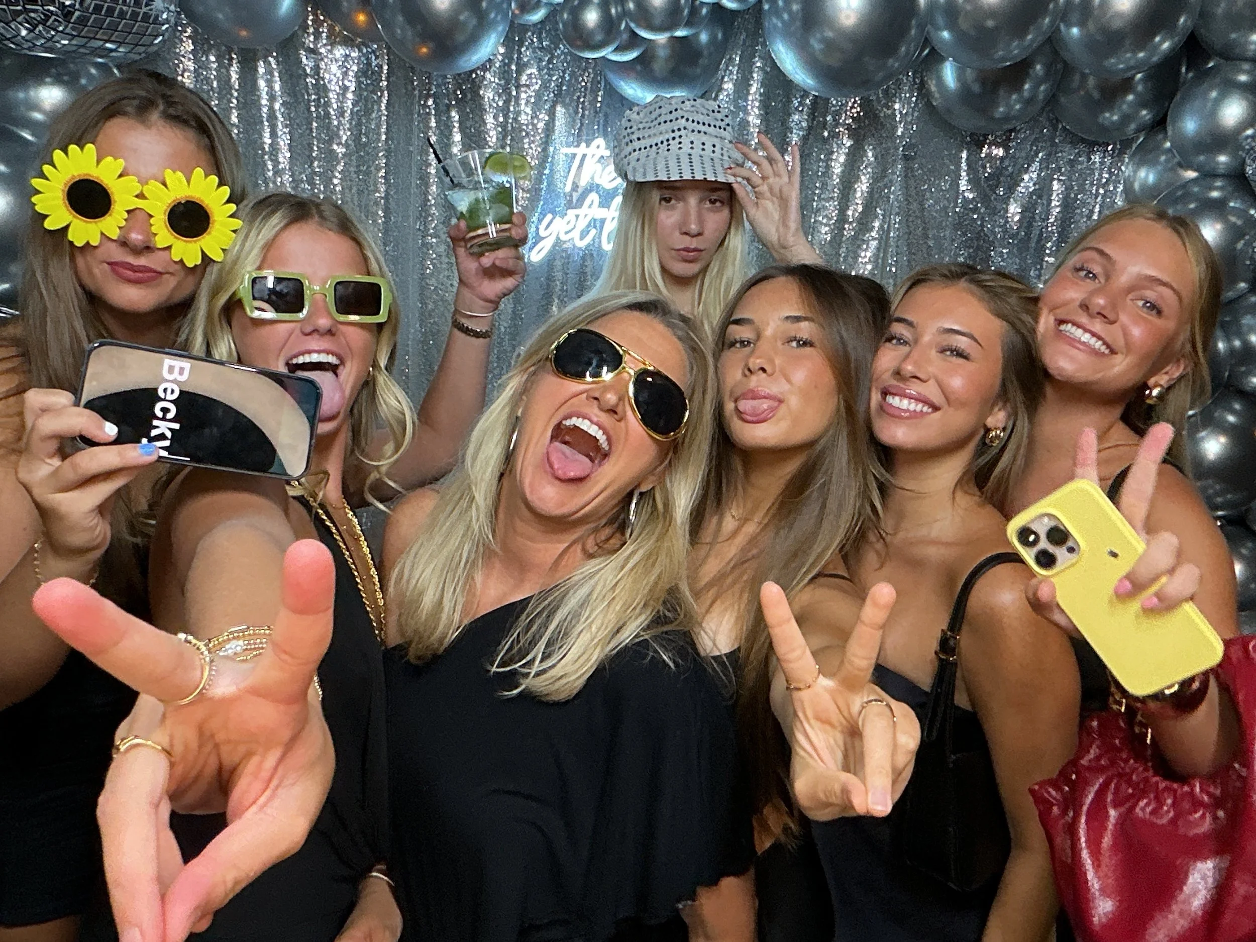A group of eight women celebrating at a party, smiling, and making peace signs. They are wearing fun accessories like sunflower glasses, sunglasses, and a hat. Some are holding drinks and a yellow smartphone, with a silver balloon backdrop and a neon