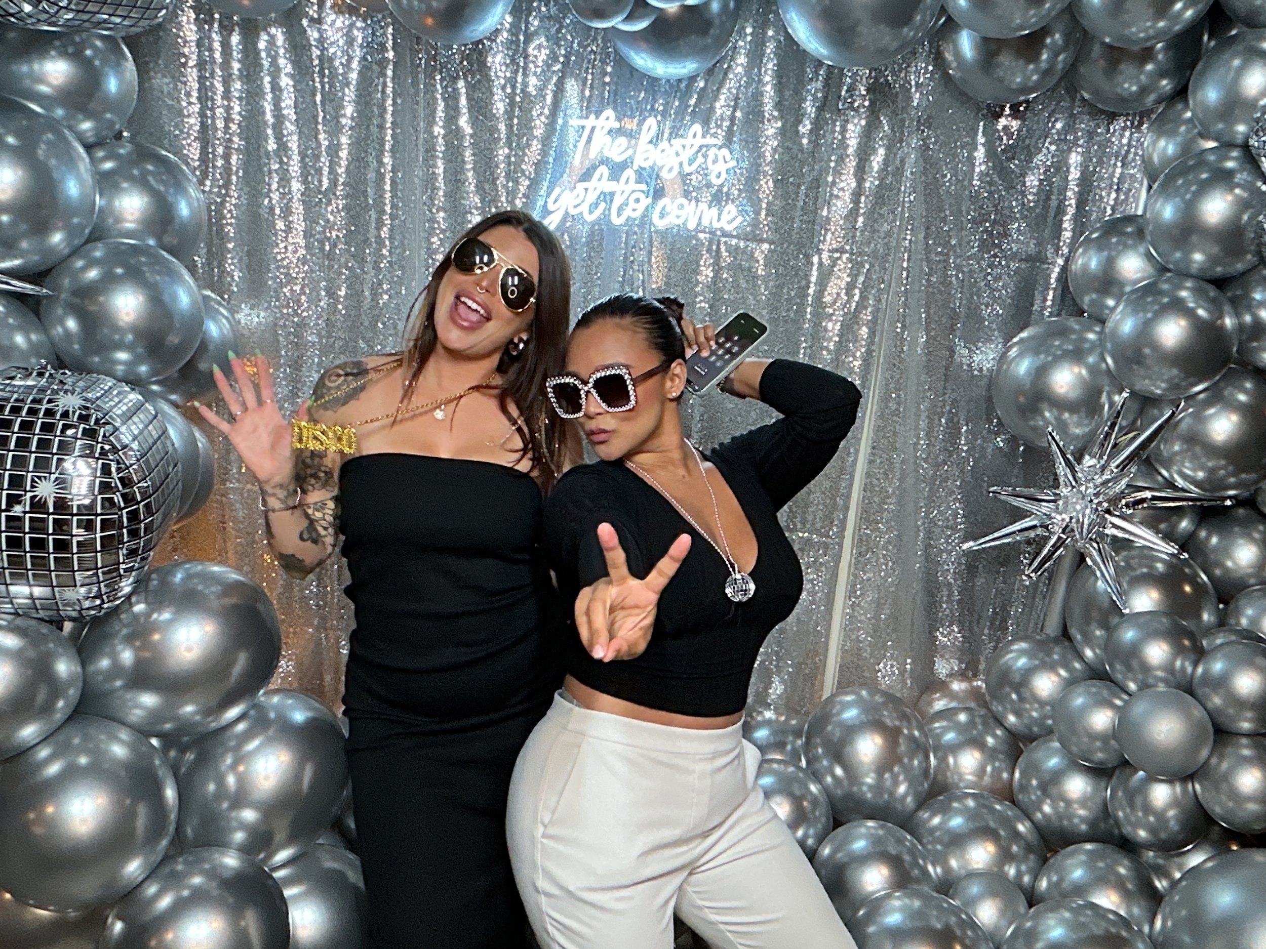 Two women dressed in black tops and light-colored bottoms posing in front of a silver backdrop with balloons and a neon sign that reads "The best is yet to come." Both women are wearing sunglasses and making peace signs, with party accessories.