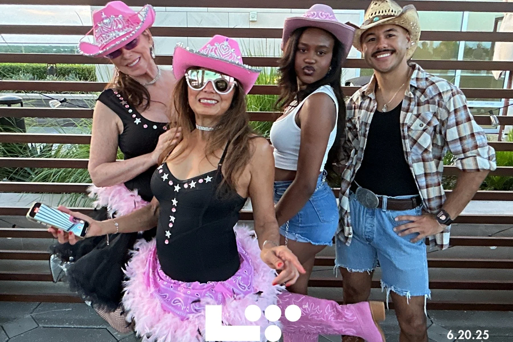 Four people dressed in western-themed costumes, wearing cowboy hats and accessories, standing outdoors near a wooden fence, smiling and posing for the photo.