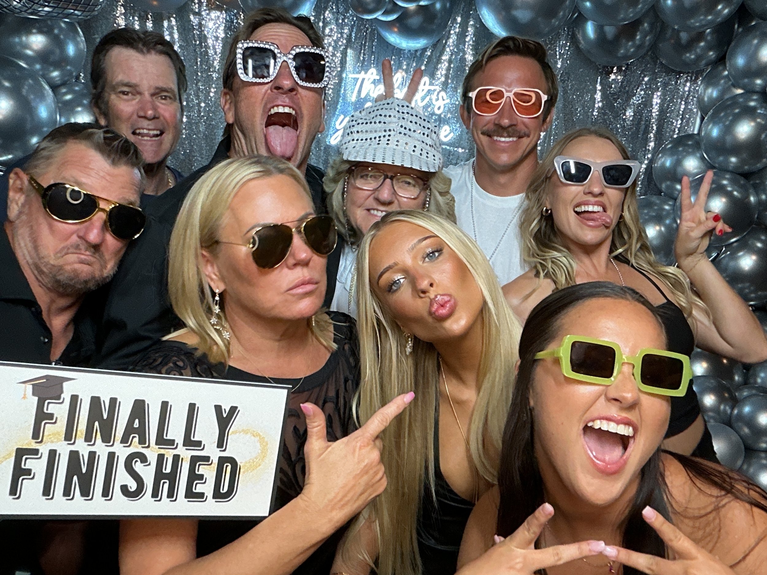 Group of eight people celebrating a graduation, wearing novelty sunglasses, with a backdrop of metallic balloons and a sign that says 'Finally Finished.'