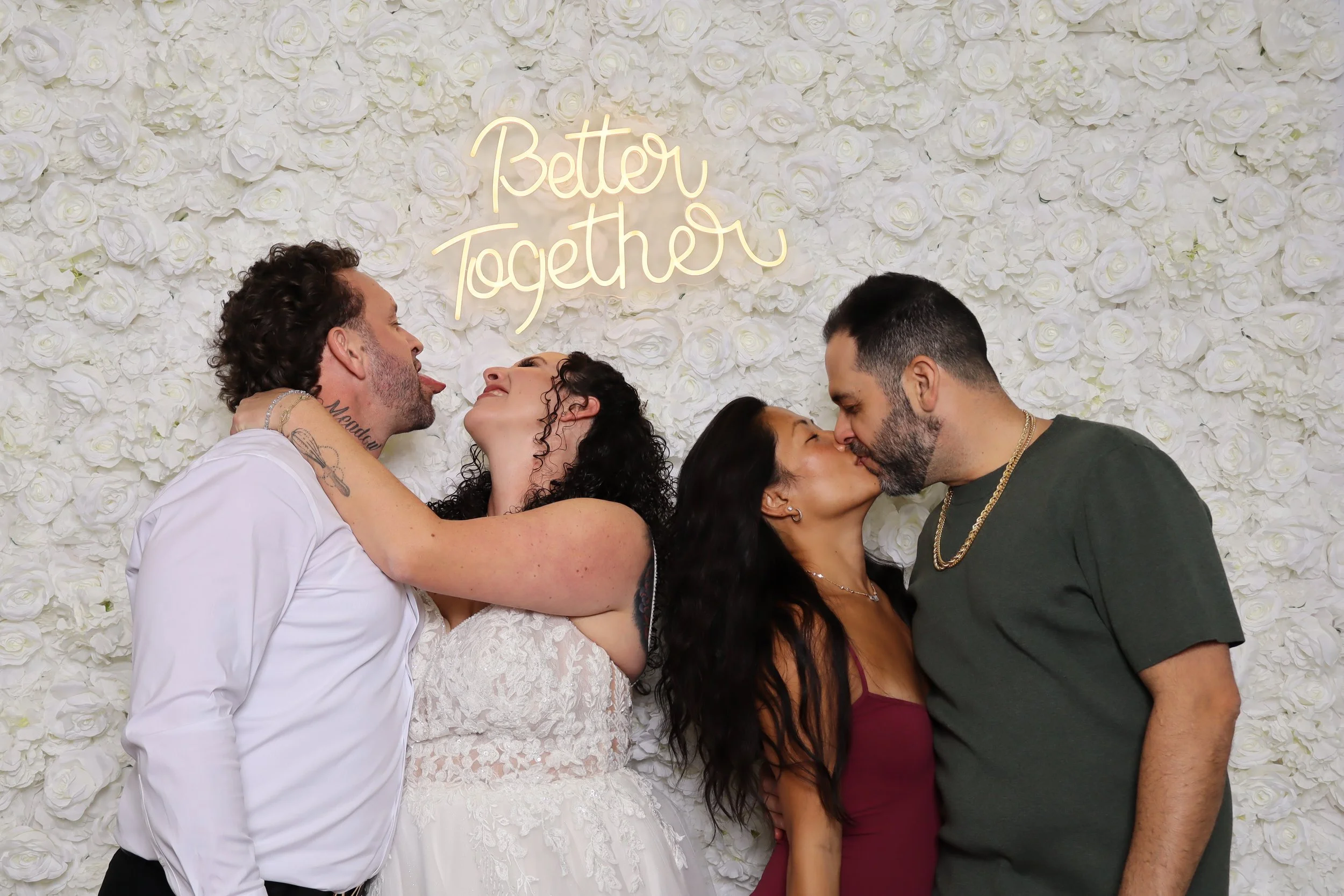 Four people kissing each other, with two men and two women, standing against a white floral backdrop with a neon sign that reads "Better Together."