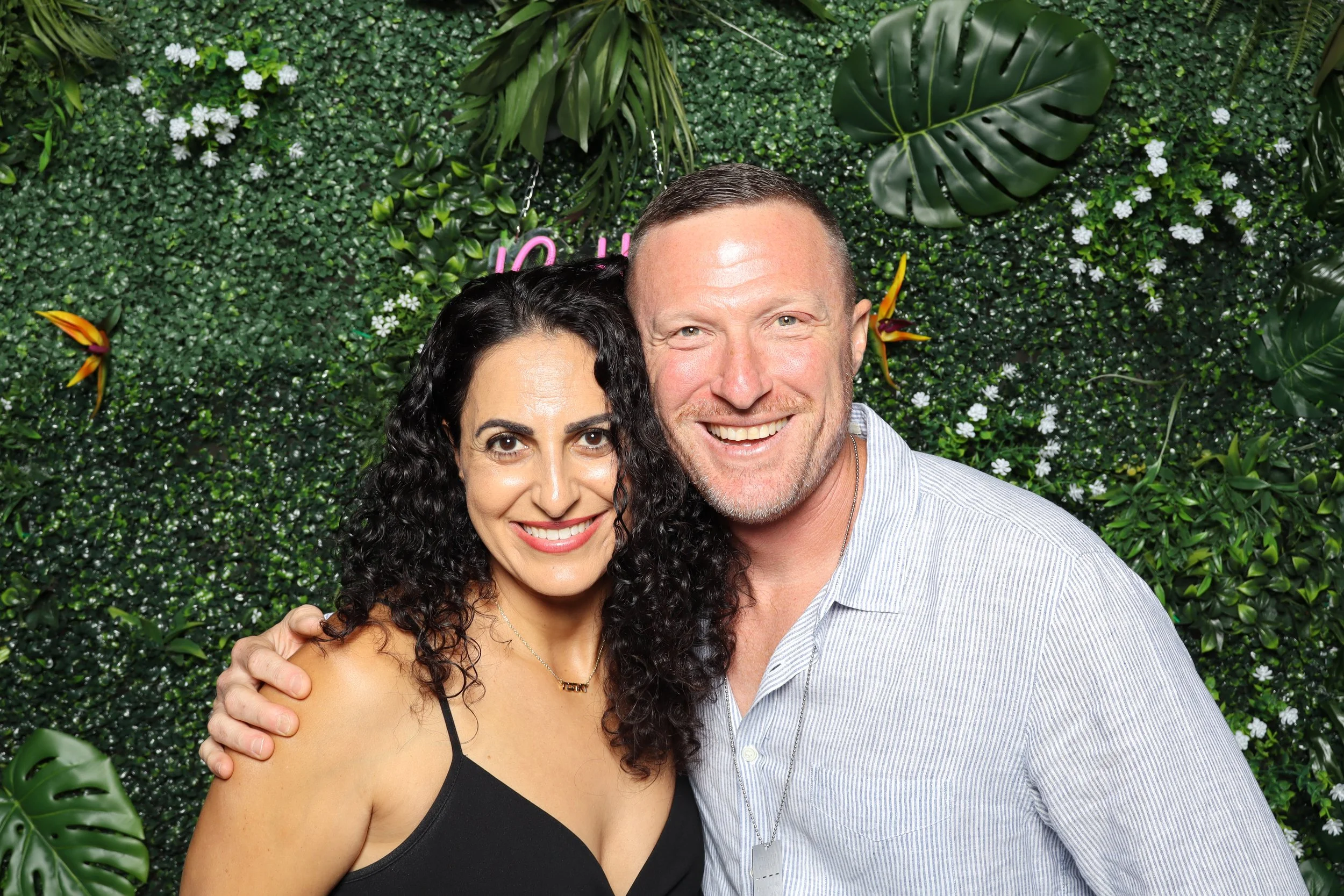 A smiling woman with dark curly hair in a black dress and a smiling man with short hair and a beard in a striped shirt, standing close together in front of a green floral wall background.