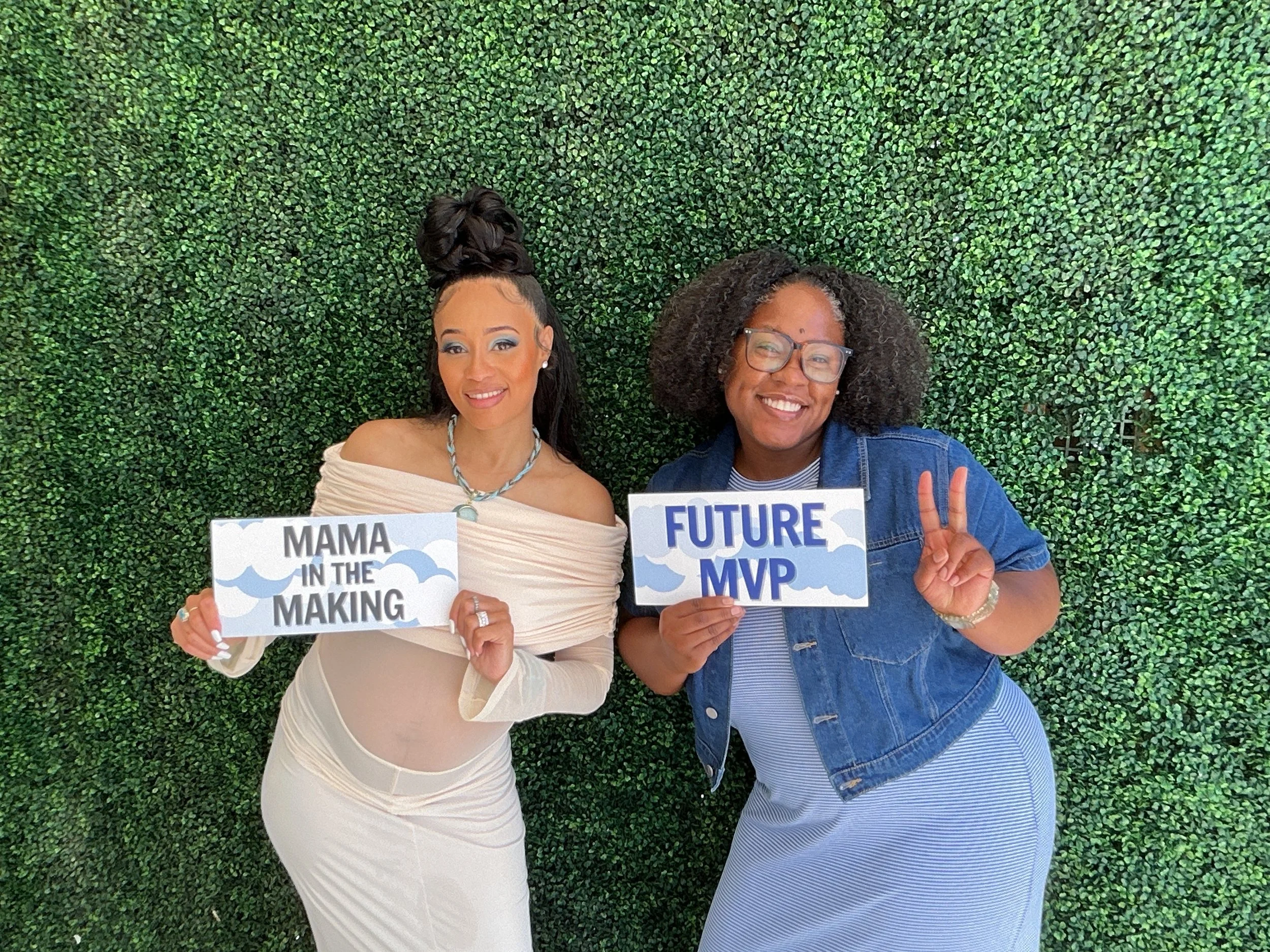 Two women standing in front of a green hedge, holding signs that say "Mama in the making" and "Future MVP," smiling and making peace signs.
