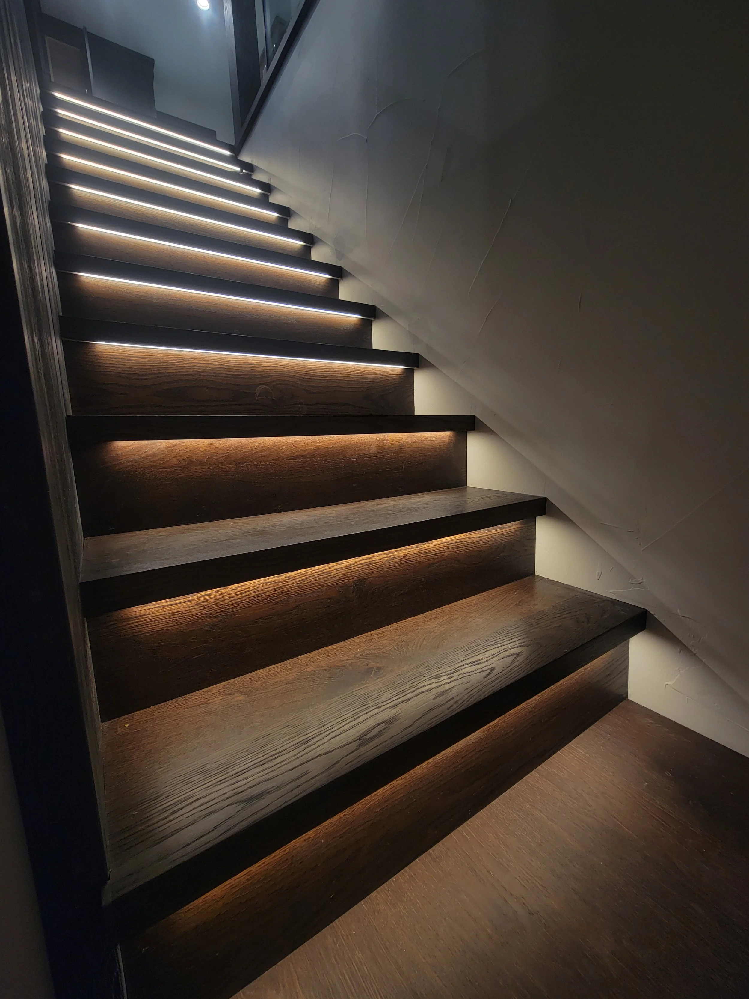 Wooden staircase with integrated LED lighting along the edges of each step, ascending towards a glass door or window at the top.