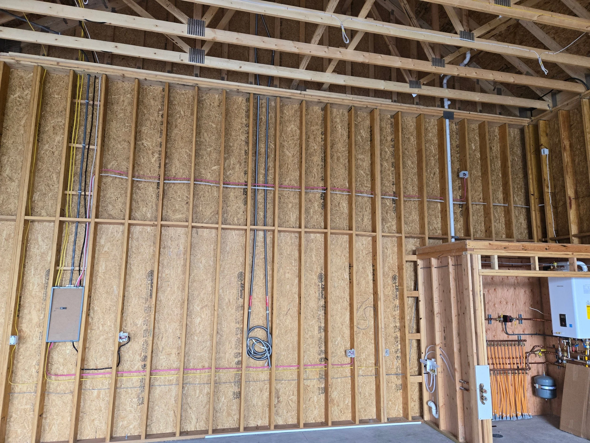 Interior view of a constructed wooden wall with exposed insulation, electrical wiring, electrical boxes, and plumbing pipes, indicating a building under construction.