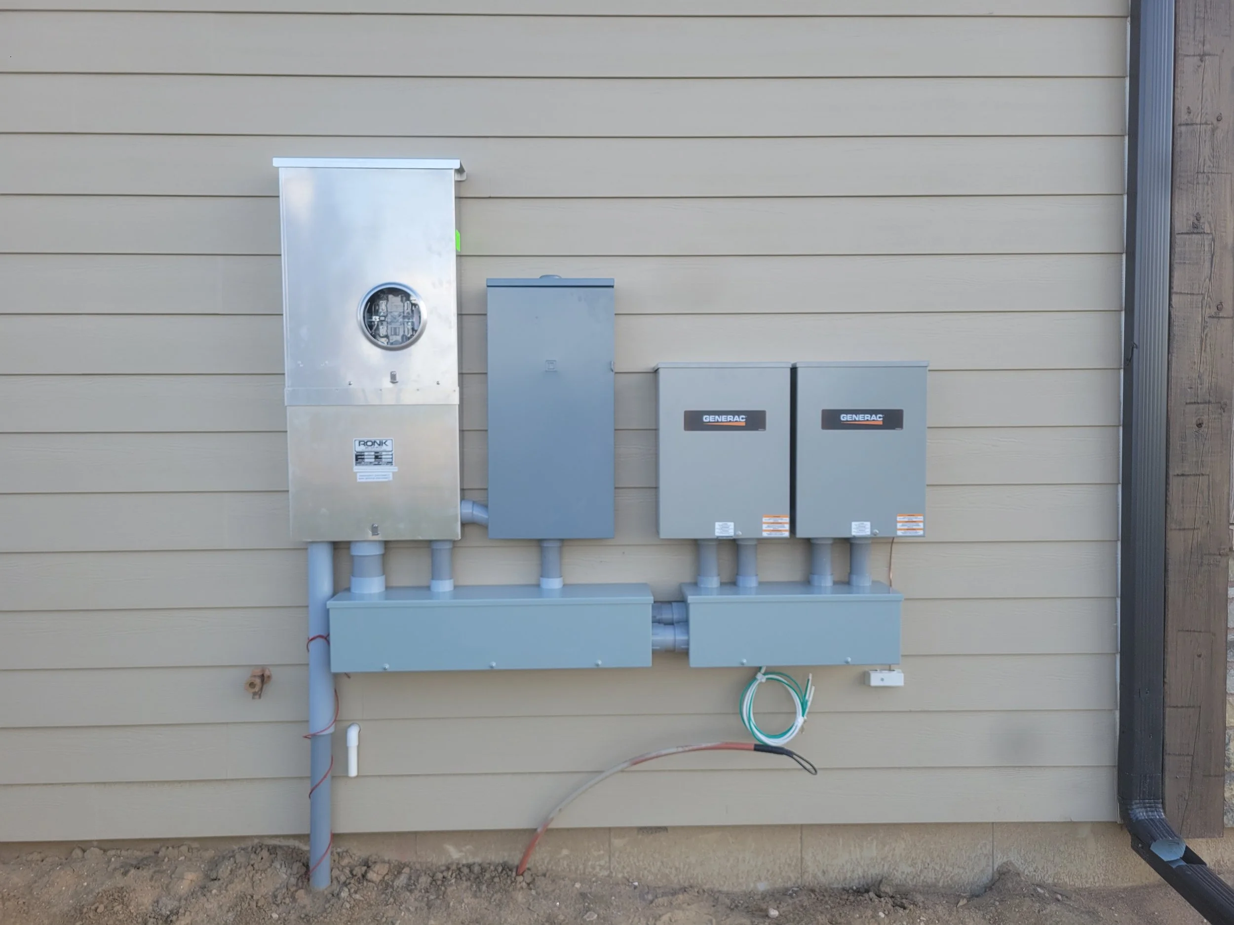 Electrical meters and panels mounted on a beige house exterior wall, including a main meter, disconnect boxes, and wiring conduit.