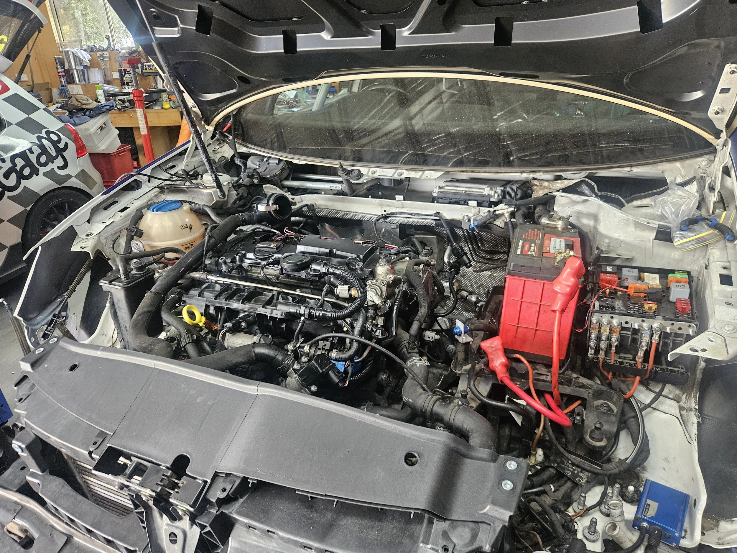 Open car hood in a garage, showing engine with various hoses, wires, and components, and a red car battery.