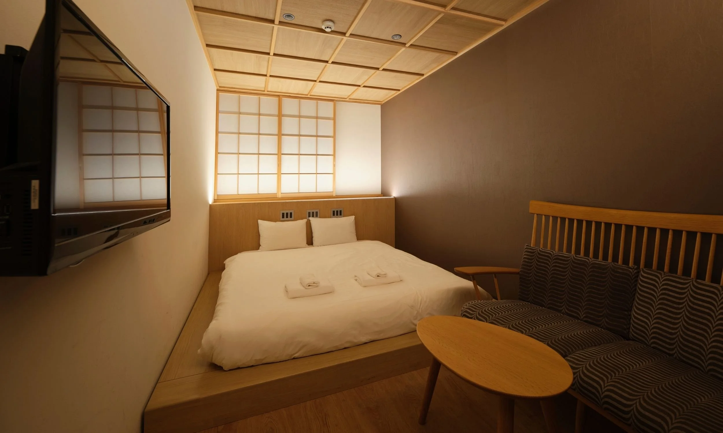 和風のデザインのモダンな客室。低いベッドには白い寝具と畳シート、枕、タオルケットが置かれている。側には木製のソファとコーヒーテーブル。壁にはスクリーン型の照明と来日したような格子窓。