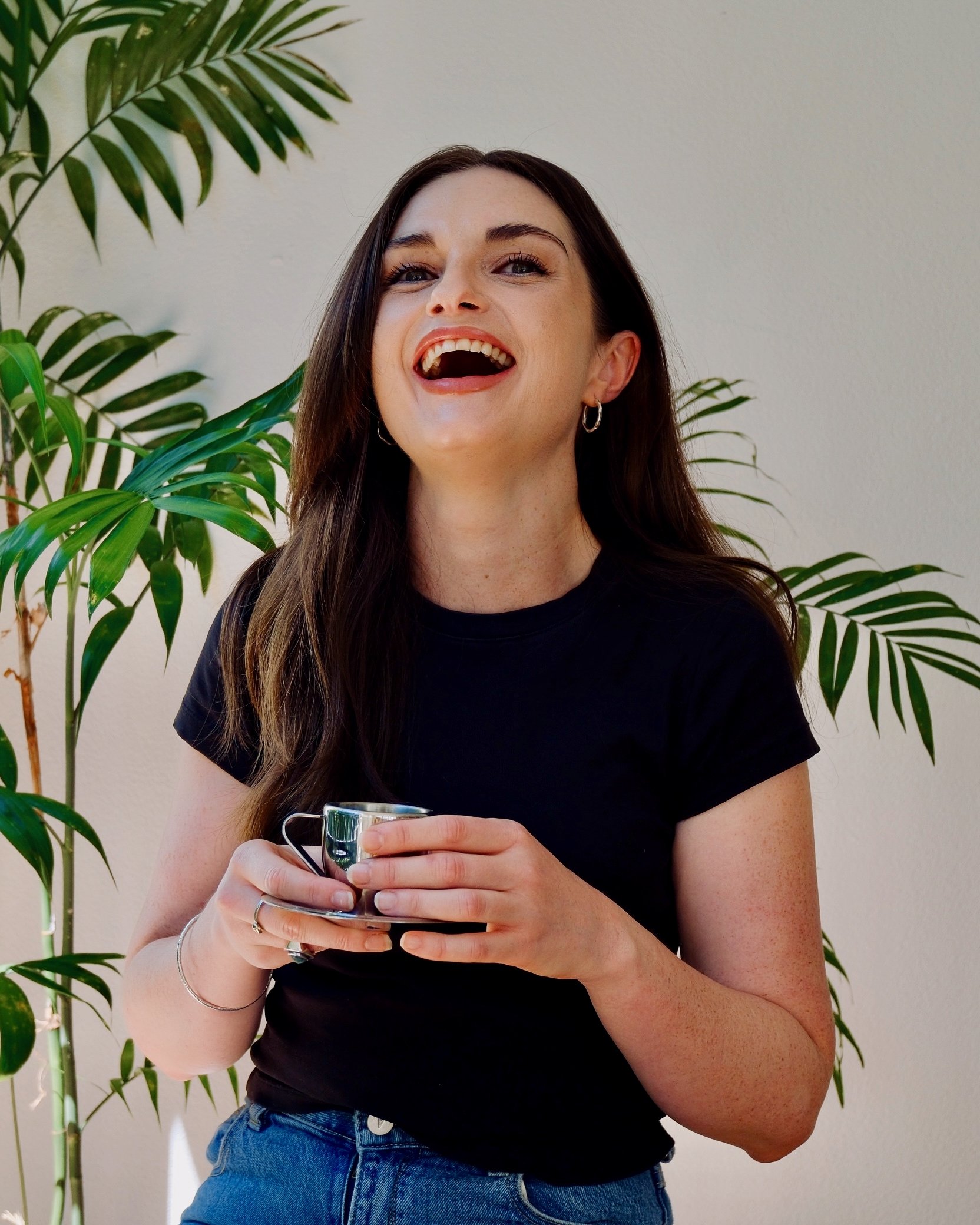 A woman with long brown hair, wearing a black shirt and hoop earrings, holds a small silver cup with both hands, standing in front of green houseplants and a plain white wall.