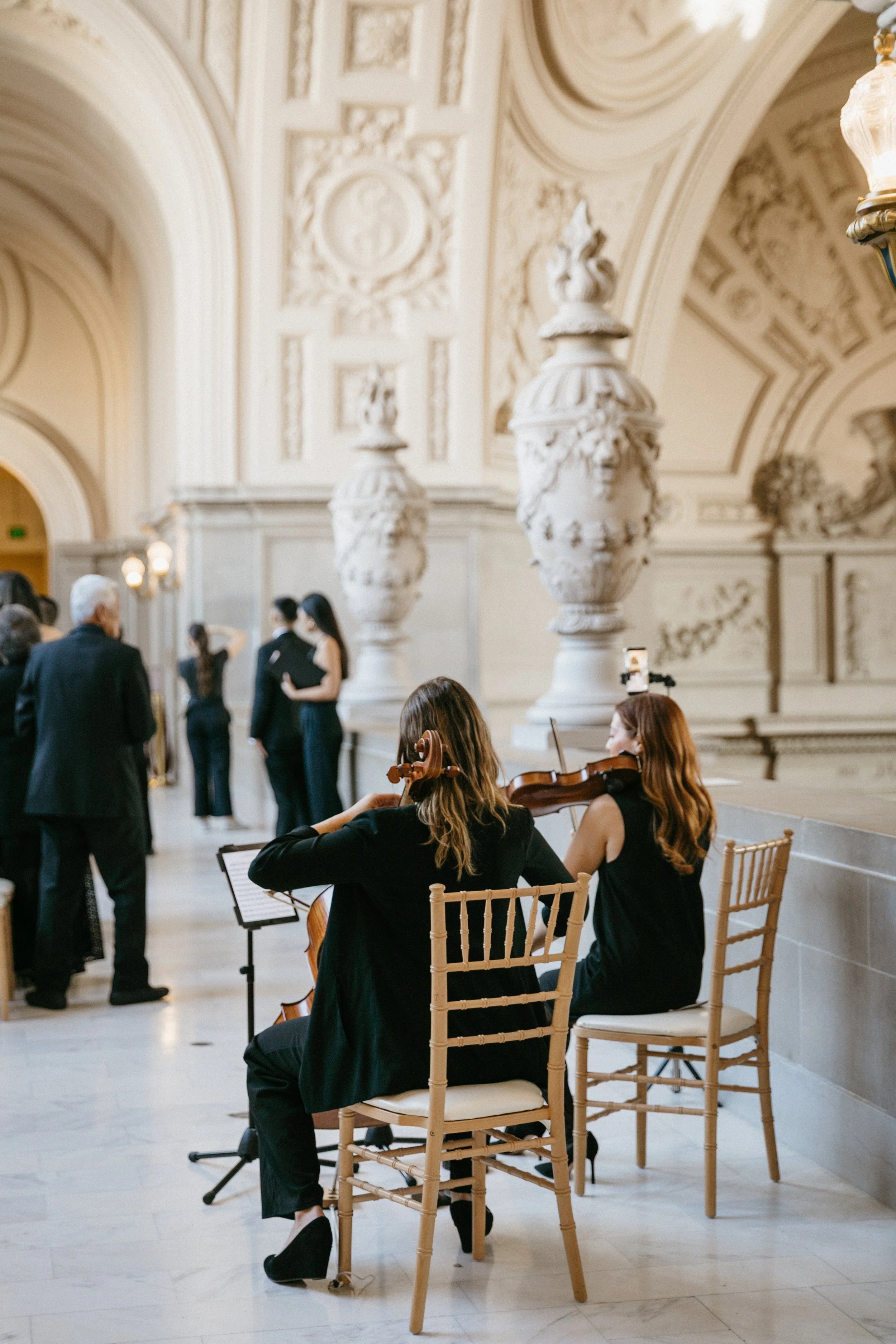 Two women playing violins in a grand, ornate hall with people in the background at SF cityhall