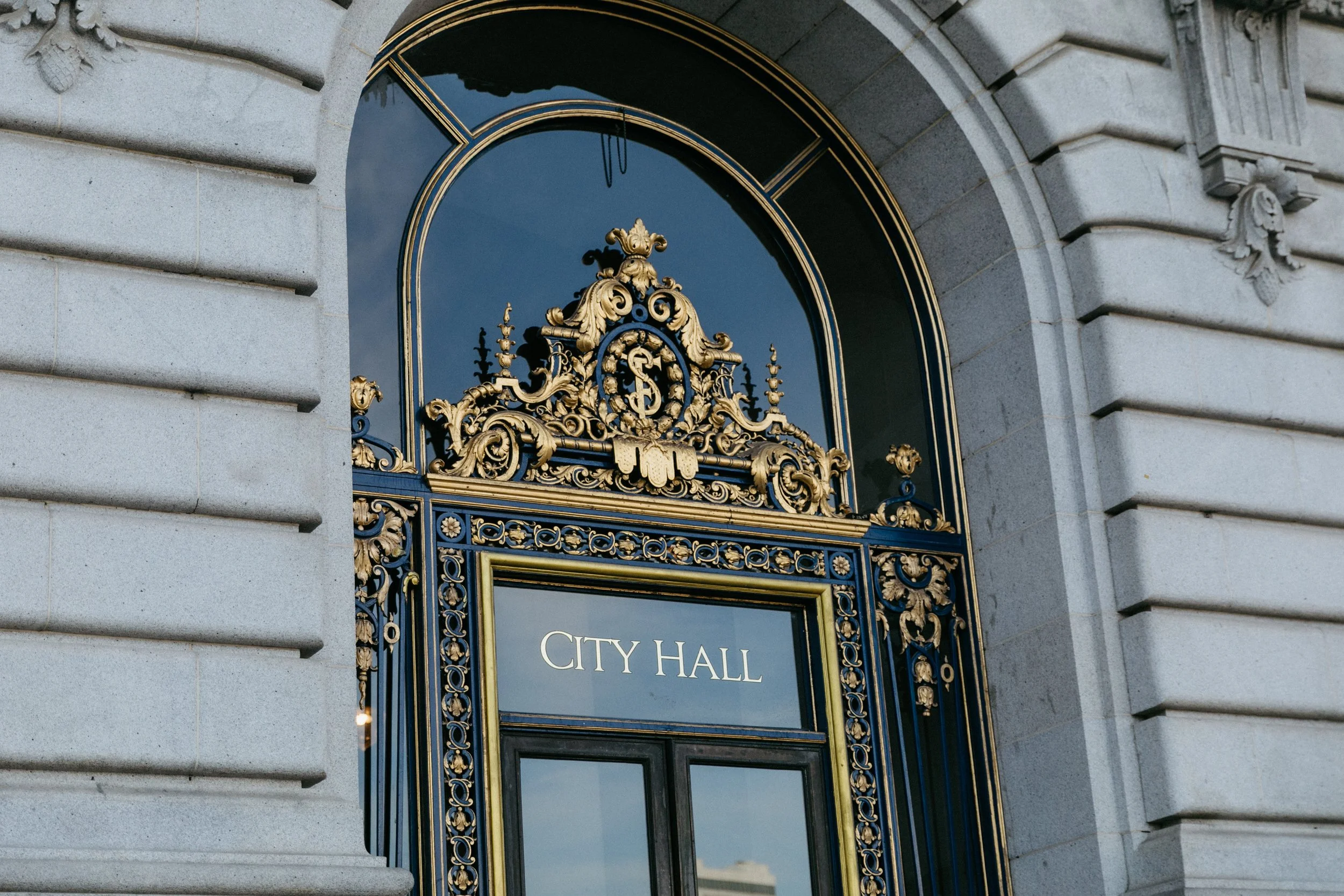 Glass door with gold ornate frame and a sign that reads 'City Hall' above the entrance. Surrounding the door is a stone facade with decorative details at sf cityhall.