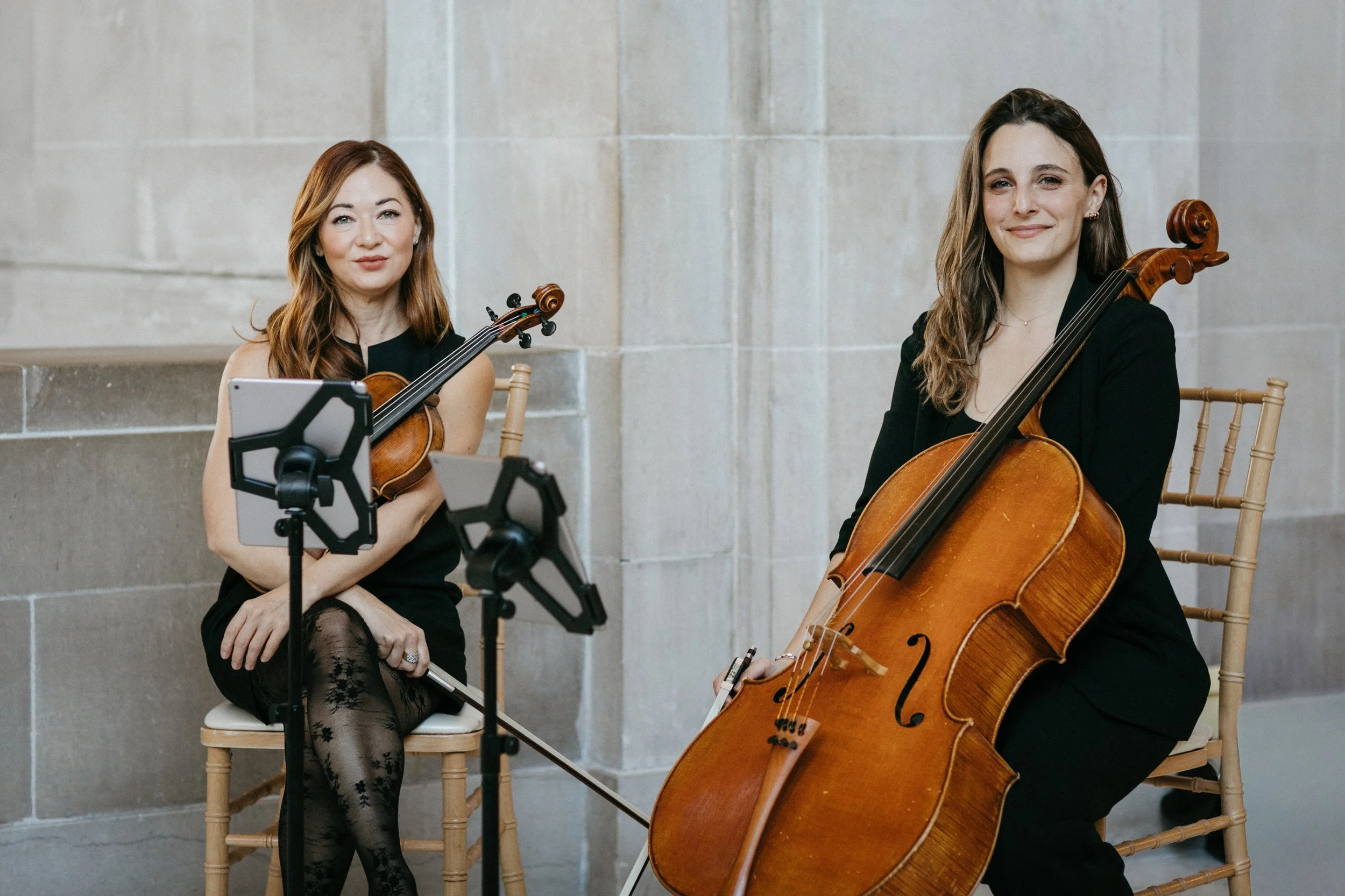 Two women sitting with string instruments, a violin and a cello, in a room with stone walls. SF CityHall. One woman has red hair and is holding a violin, while the other woman has brown hair and is holding a cello.