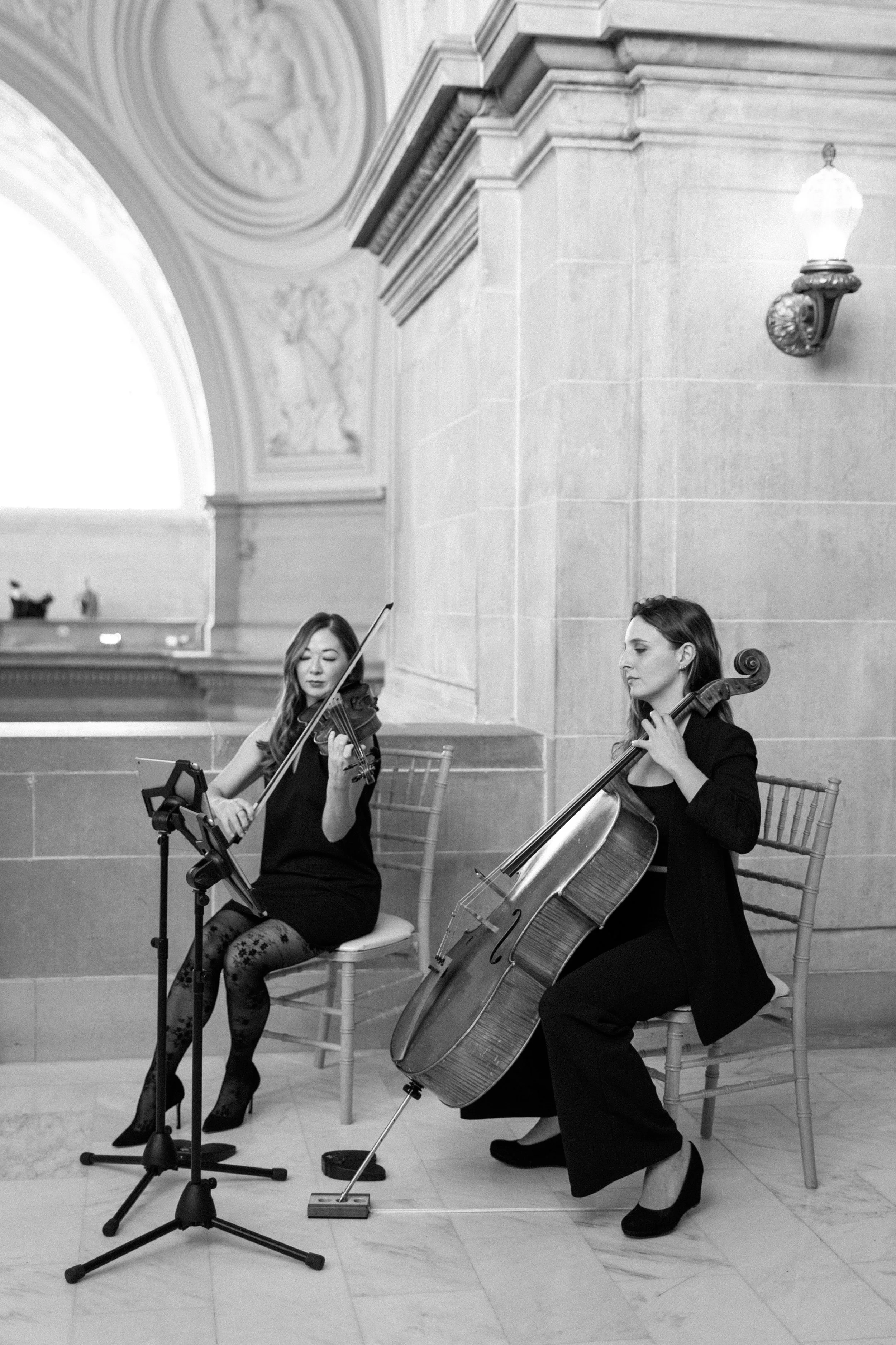Two women playing string instruments, a violin and a cello, in an elegant indoor setting with marble walls and ornate architectural details at SF Cityhall.