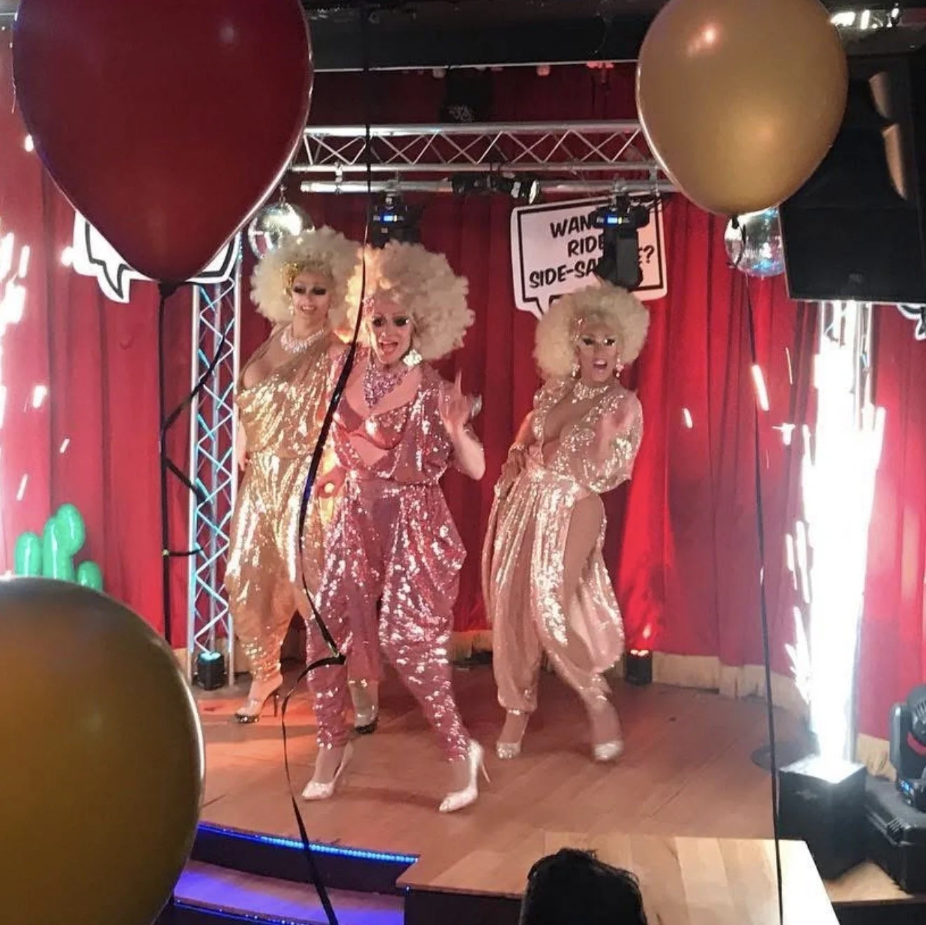 Three drag performers on stage in shiny, sequined outfits and big, curly platinum blonde wigs, dancing and singing with red curtains behind them. Balloons in the foreground and a sign in the background reads 'Want a ride? Side-Sash?'