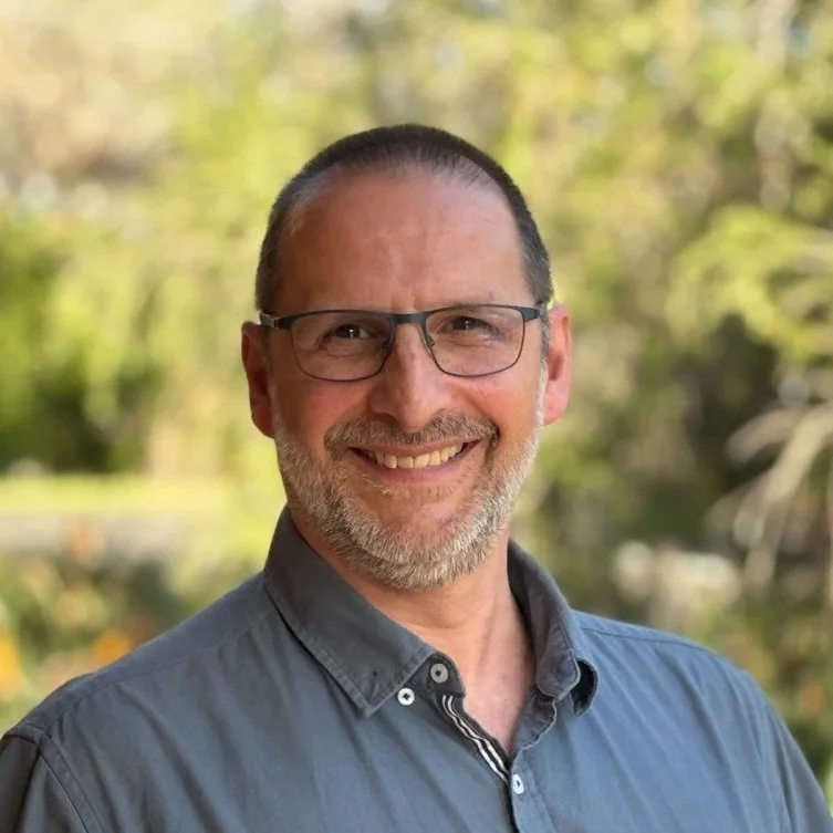 A middle-aged man with glasses, a gray beard, and short hair, smiling outdoors with a blurred background of trees and greenery.