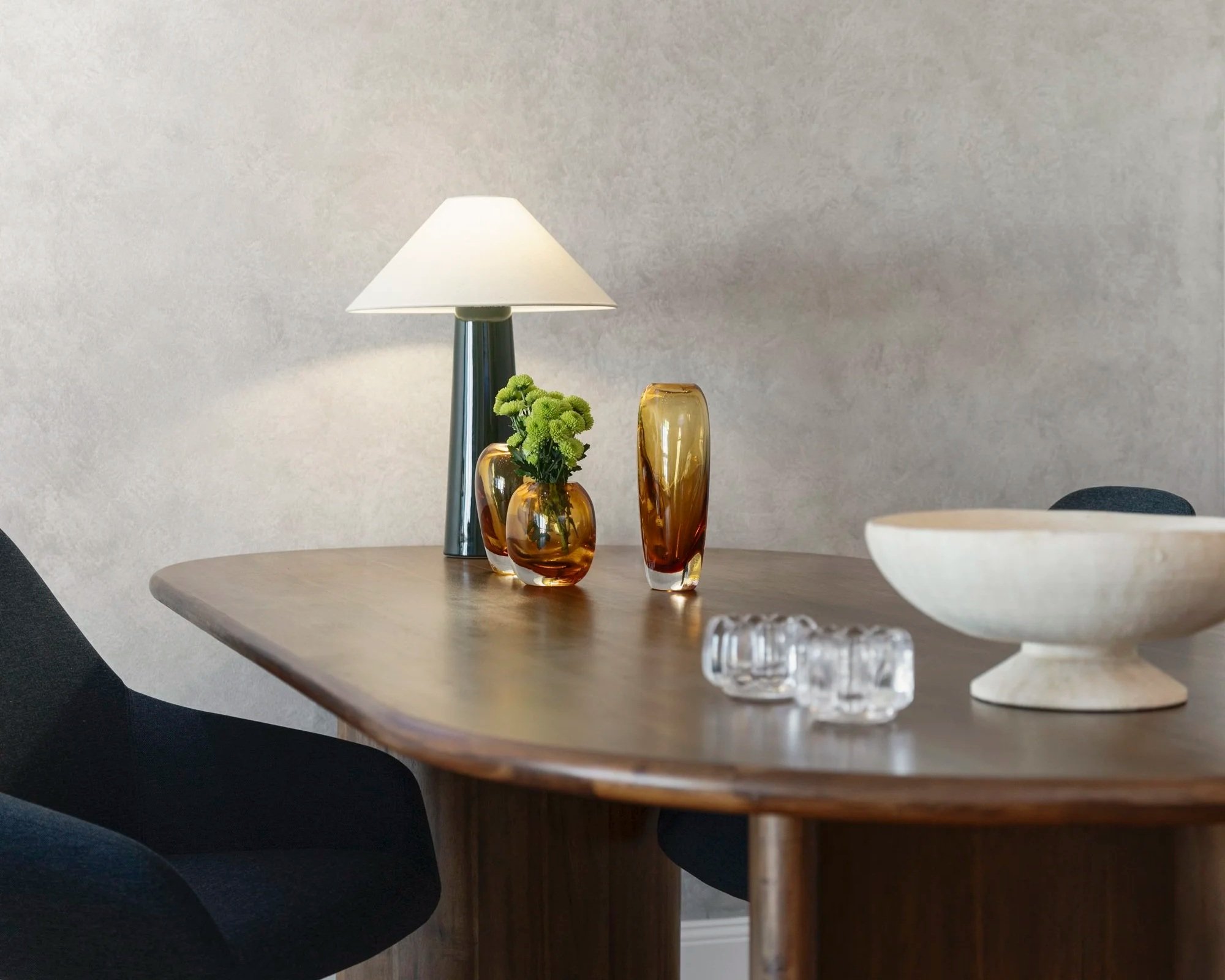 A wooden dining table with a black chair, decorated with amber vases, a white table lamp, a white ceramic bowl, and glass decorative objects, against a plain light-colored wall.