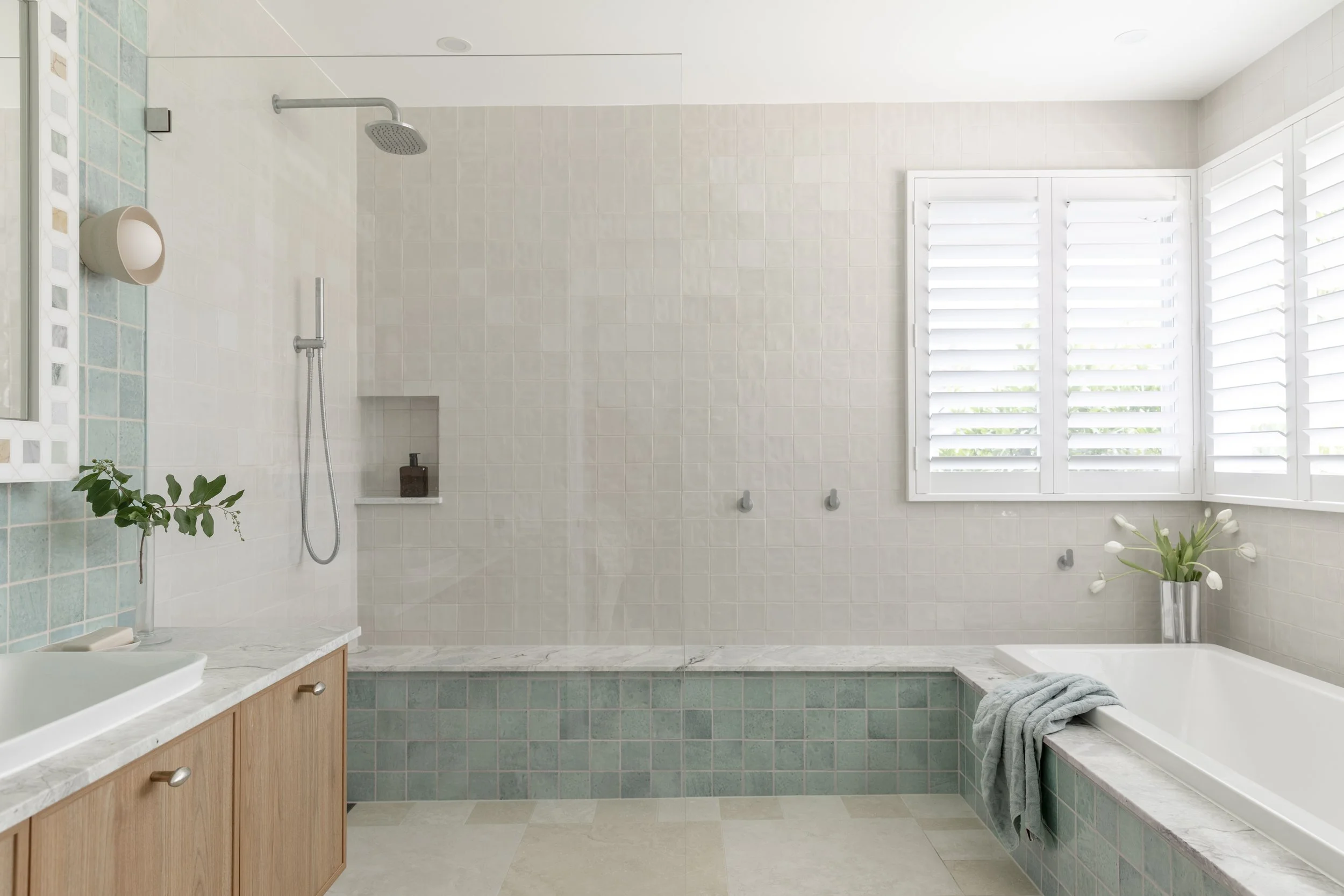 A clean, modern bathroom with a walk-in shower, a bathtub, a window with white shutters, and a wooden vanity with a marble countertop. There is a small plant on the vanity and a vase with white tulips near the bathtub.