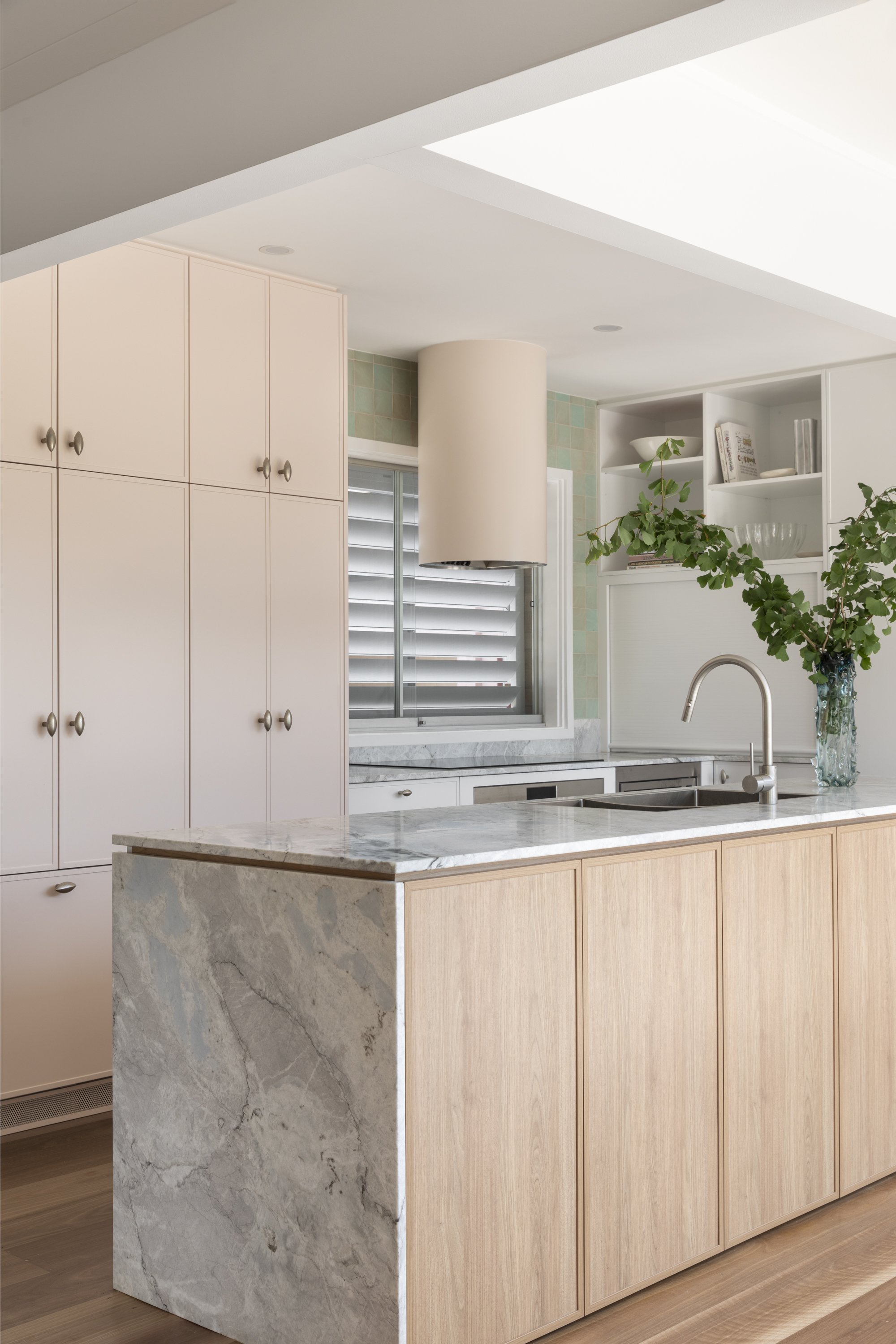A modern kitchen with white cabinetry, a marble kitchen island with wood paneling, a large window with shutters, and decorative shelving with plants and dishes.