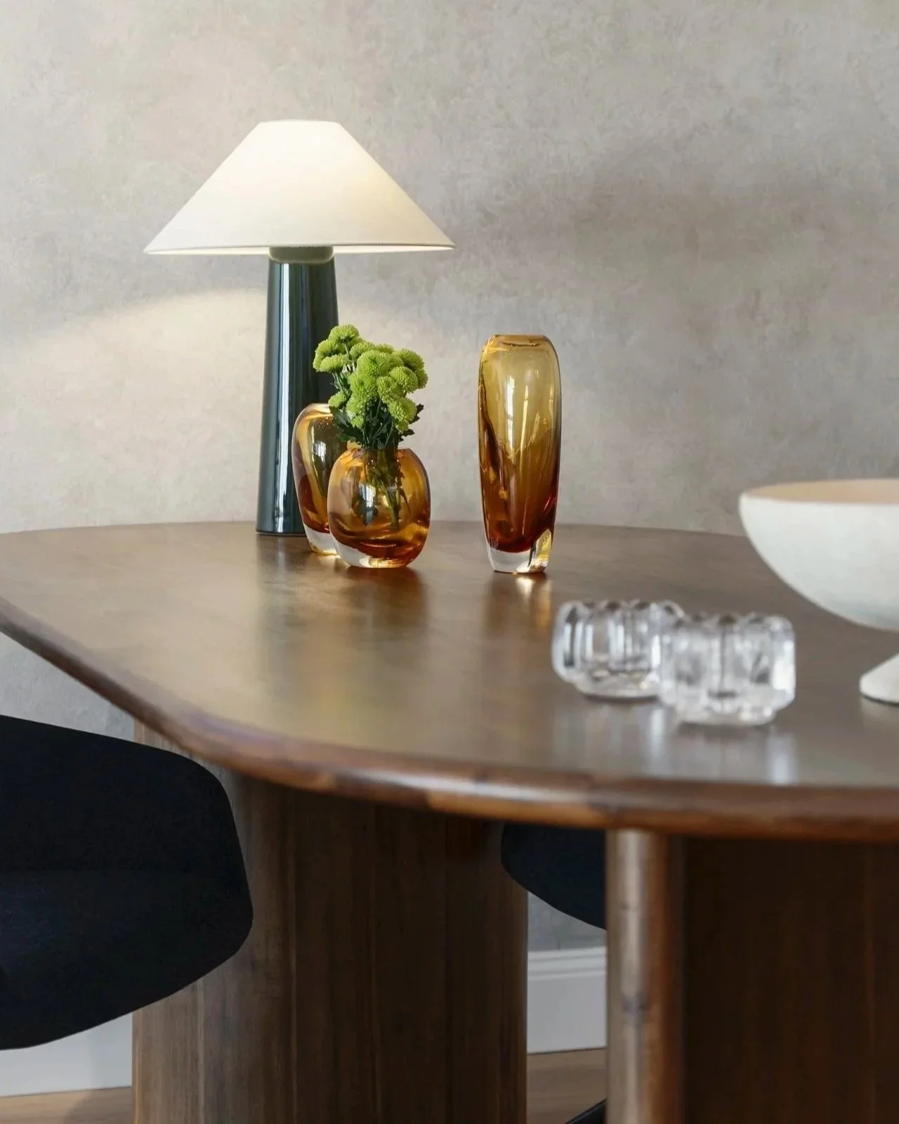 A modern dining room with a wooden table, black chairs, a table lamp, amber glass vases, and a white bowl on the table.