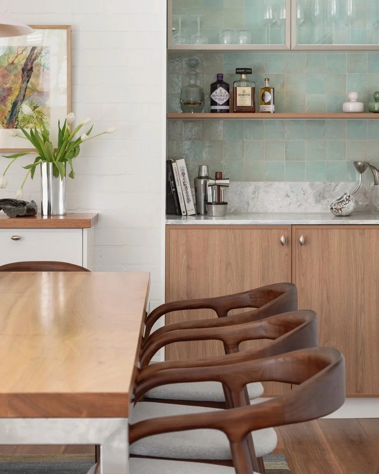 Dining room with a wood table, mid-century modern wooden chairs with light-colored cushions, a sideboard with a vase of white tulips, and a built-in glass-front cabinet with glassware and bottles.