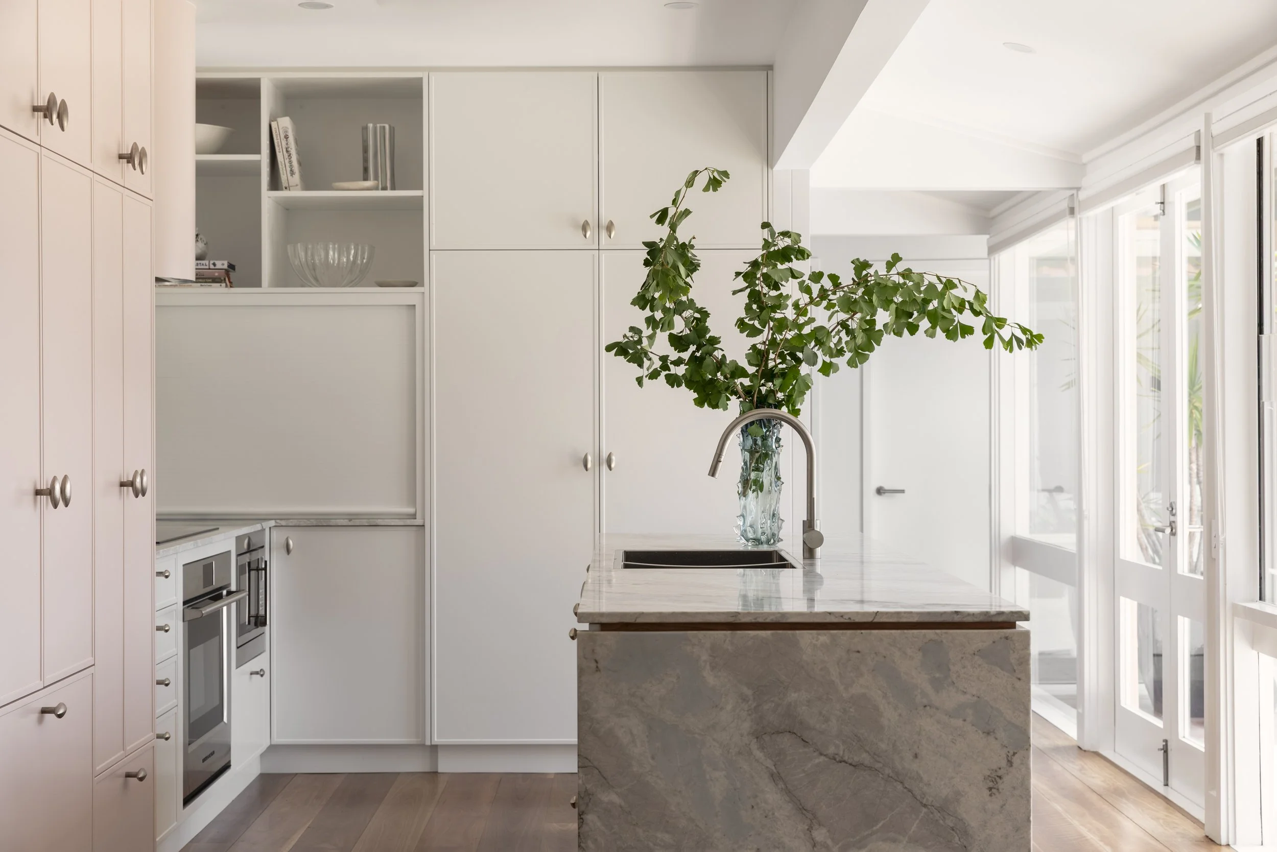 Modern kitchen with white cabinets, marble island, and a tall green plant in a glass vase on the island. Large windows with natural light.
